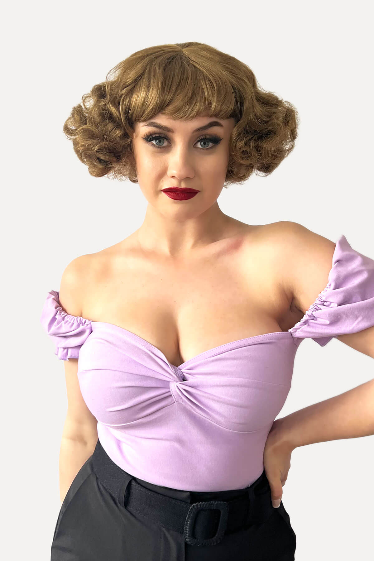 Woman wearing a light purple off-shoulder top with a white background. A short brown vintage style wig with waves: Rita golden brown 12-27