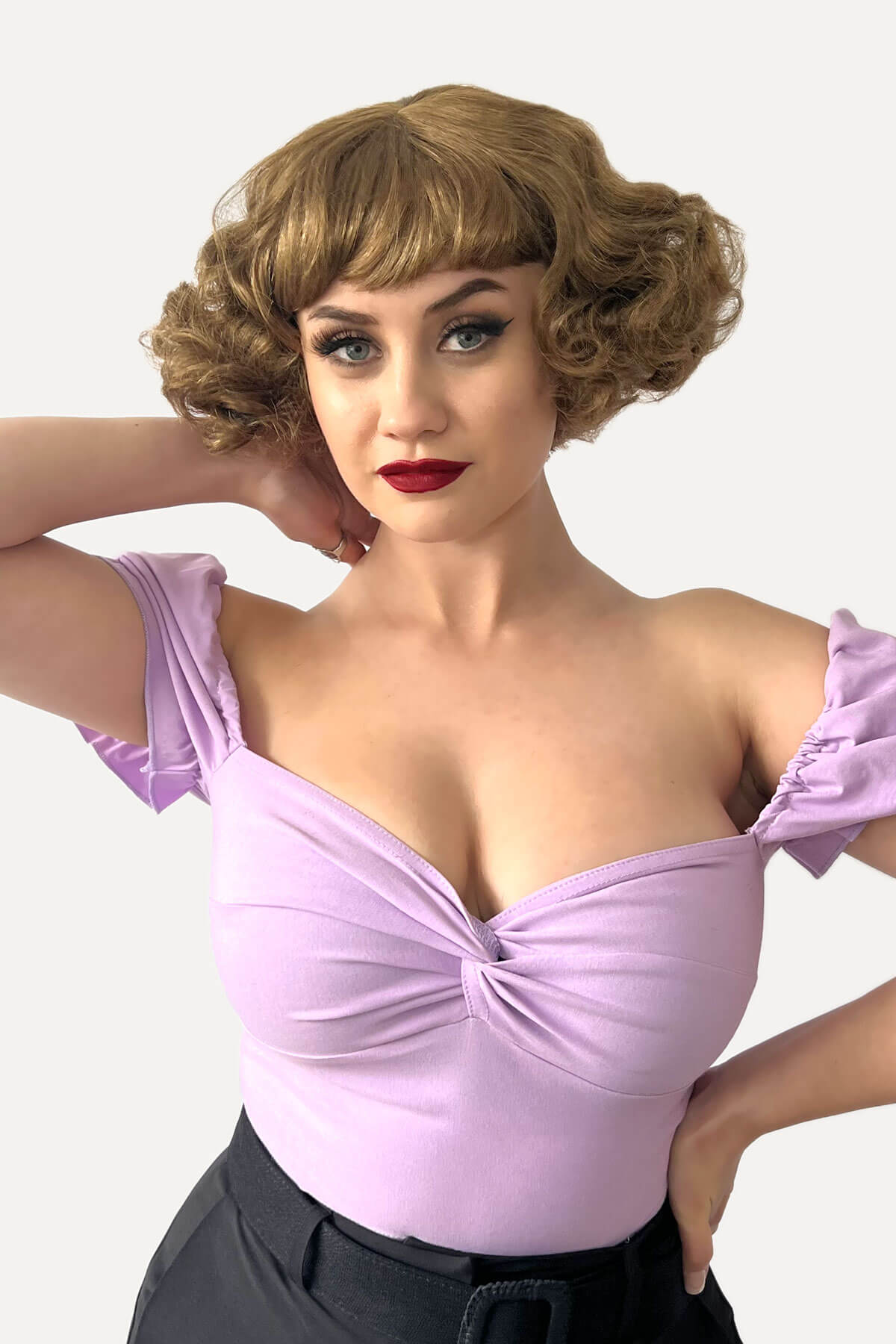Woman wearing a light purple top with puffed sleeves against a white background. A short brown vintage style wig with waves: Rita golden brown 12-27