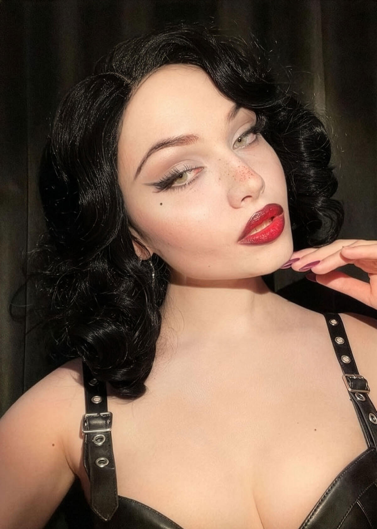Woman with dark hair and red lipstick wearing a black leather outfit against a dark background. Black lacefront wig, pinup/vintage style, mid length with finger waves: Etta