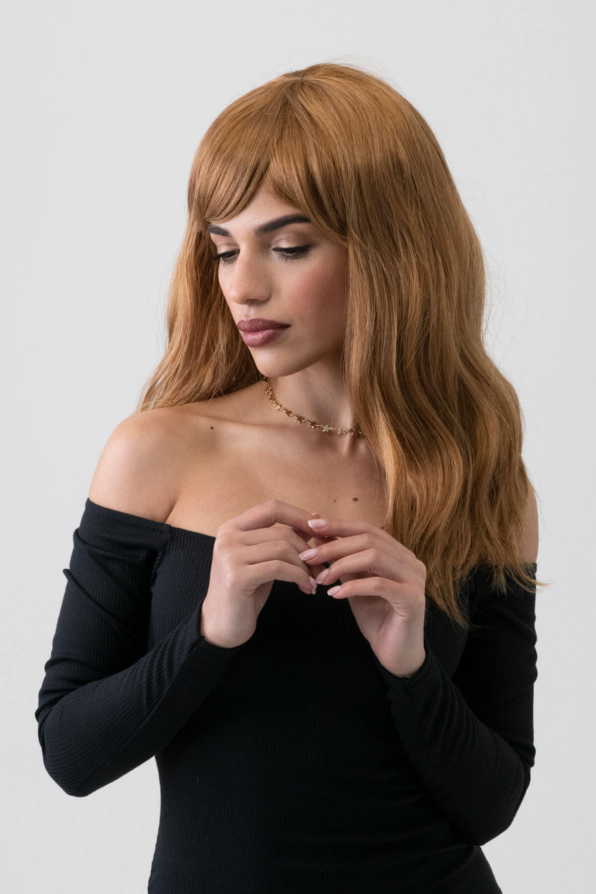 Woman with long brown hair wearing a black off-shoulder top against a white background. Golden brown wig with long waves: Carmella