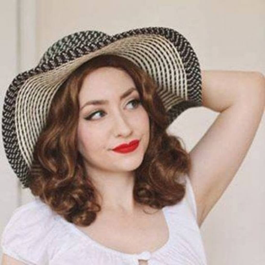 Woman wearing a wide-brimmed straw hat with a neutral background. Brown 1940s pinup style wig, with finger waves: Honey light brown 12