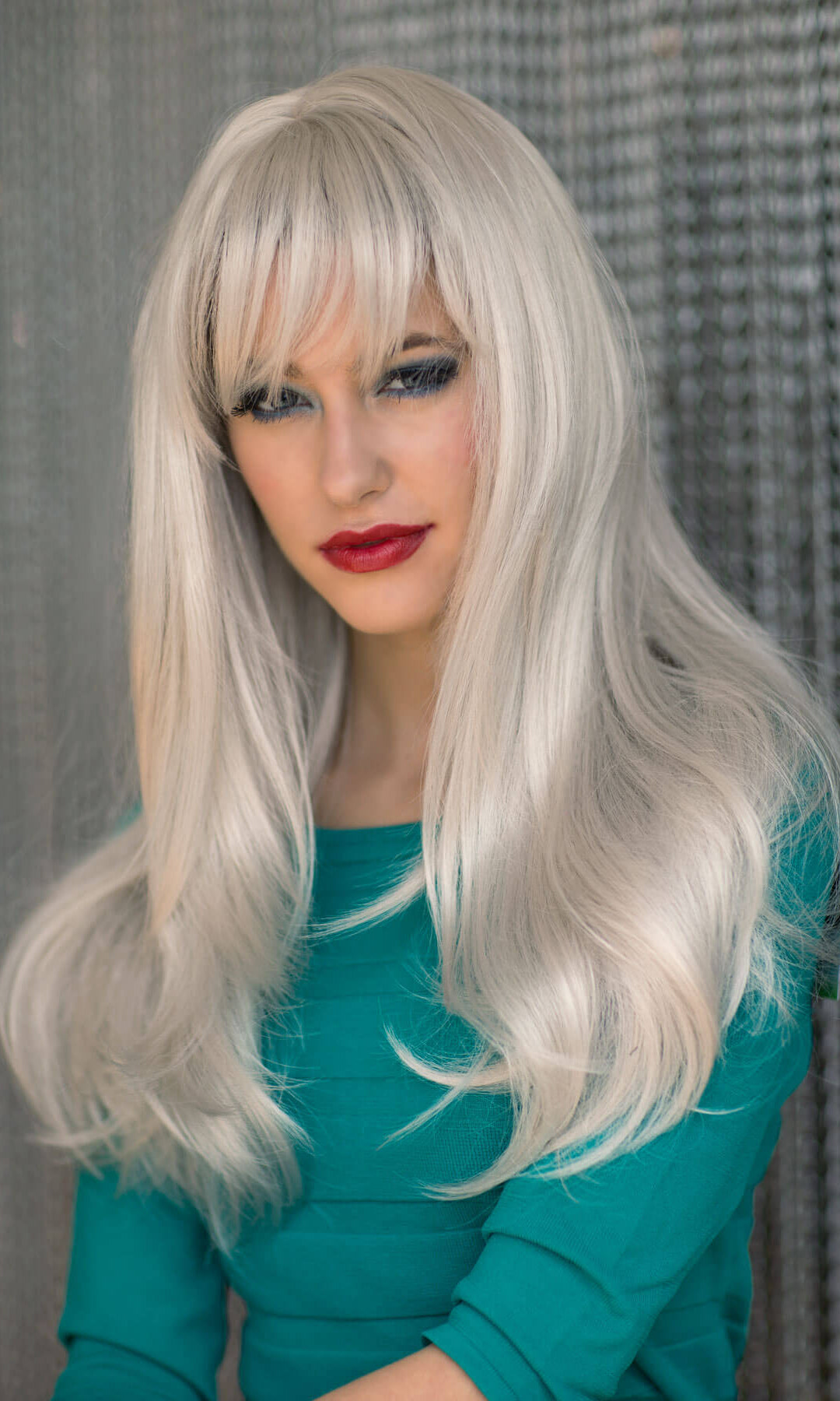 Person with long blonde wig and teal top against a textured grey background. Long silver/grey wig in razor cut, face framing style: London