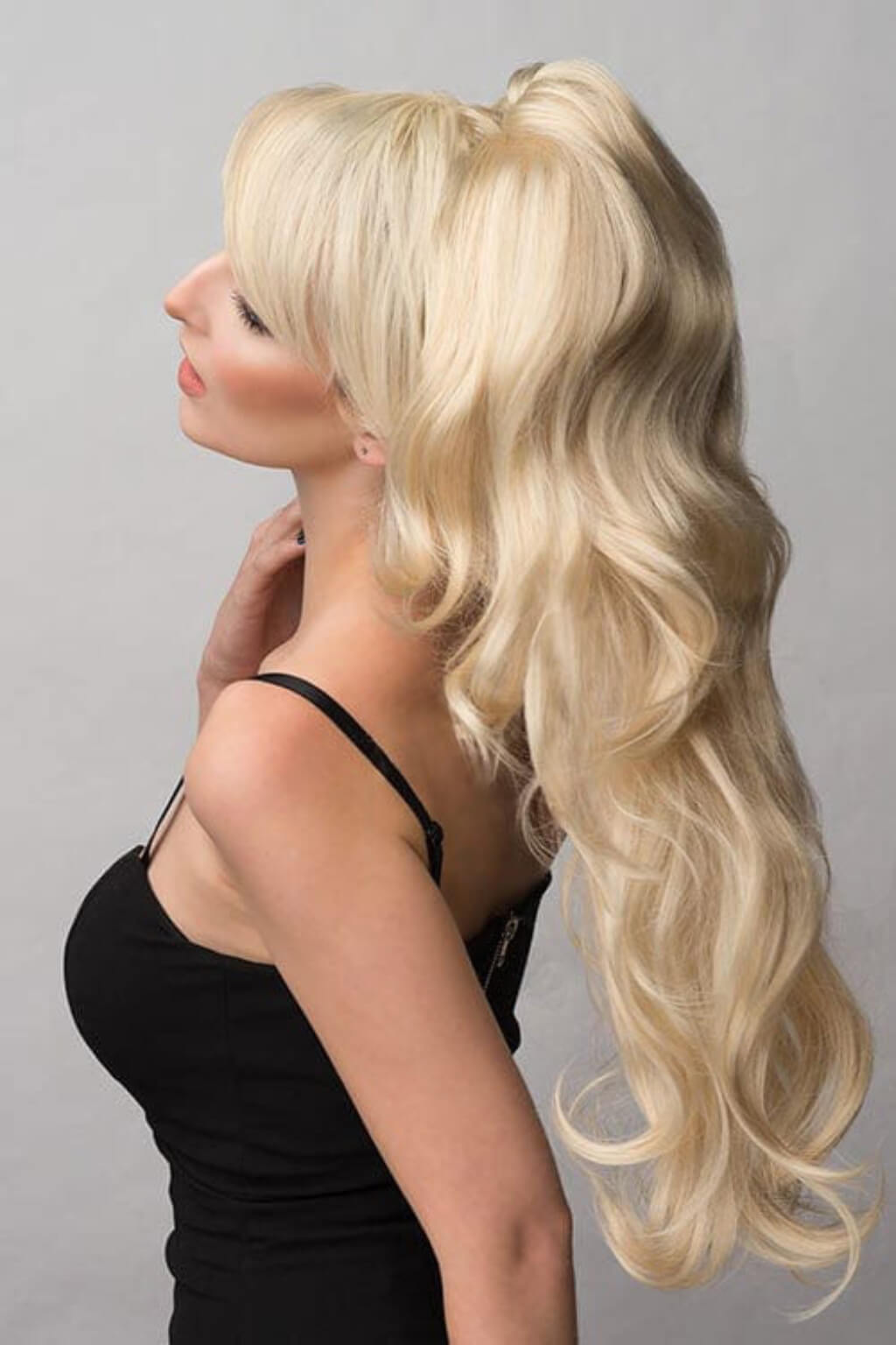 A woman shown in side profile wearing a long, wavy blonde wig with a high ponytail, photographed against a plain light grey background. Double volume hairpiece extension with gentle waves: Naomi blonde 613T24