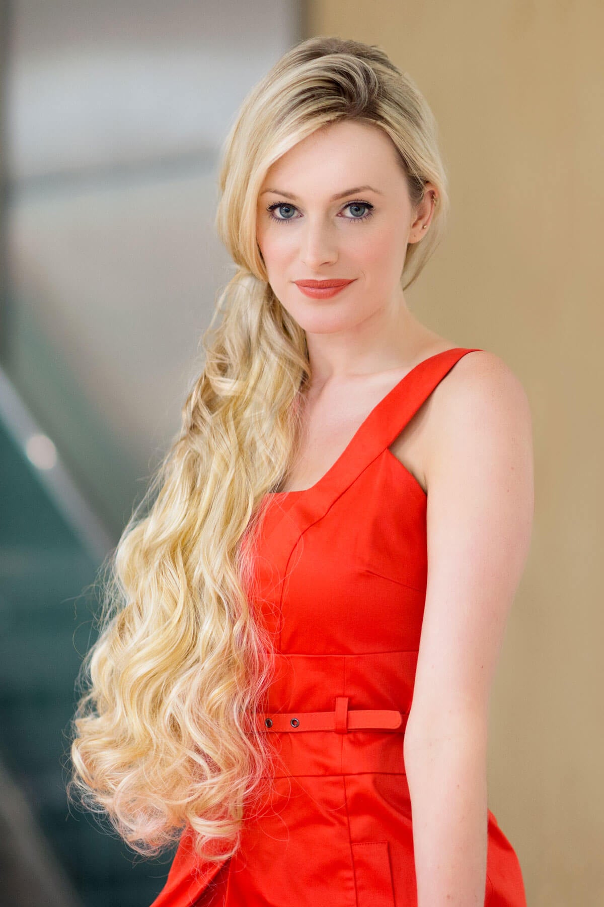 Woman in a red dress with long blonde hair against a blurred indoor background. Extension hairpiece, long, versatile, Flexihair: Cristina honey blonde 24BH613