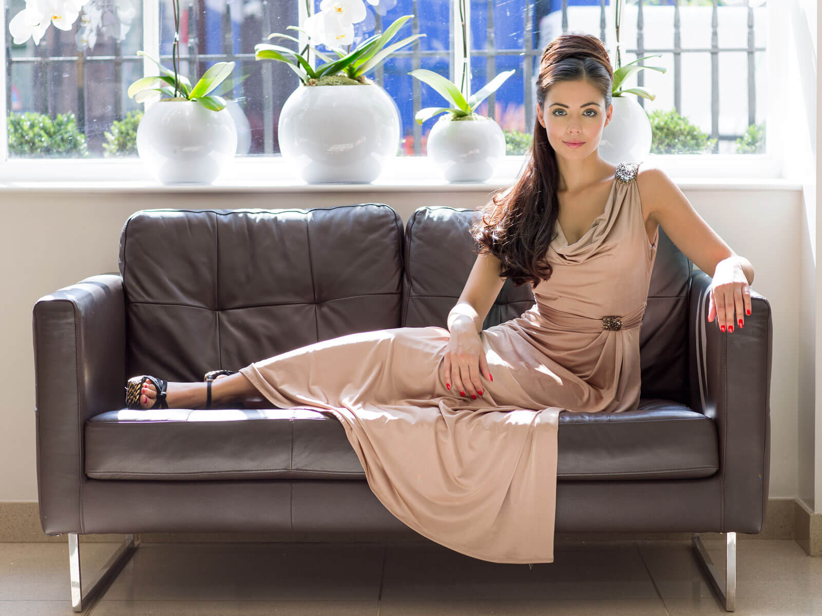 Woman in a beige dress sitting on a brown leather couch with decorative plants in the background. Extension hairpiece, long, versatile, Flexihair: Cristina chestnut brown 8
