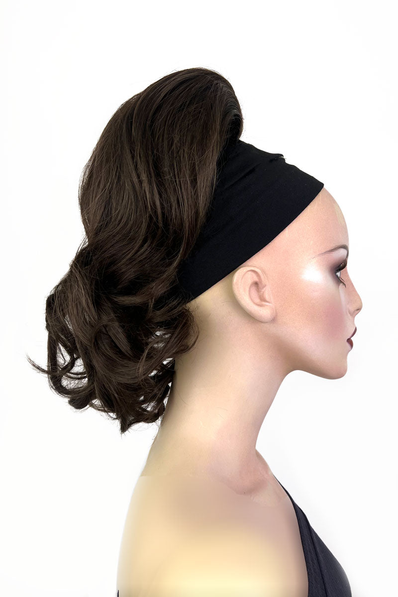 A woman wearing a curly ponytail extension hairpiece with loose, layered curls. She is seen from the shoulders up, with a backdrop featuring bokeh lights. Curly ponytail extension clip-on hairpiece: Tulisa chestnut brown 8