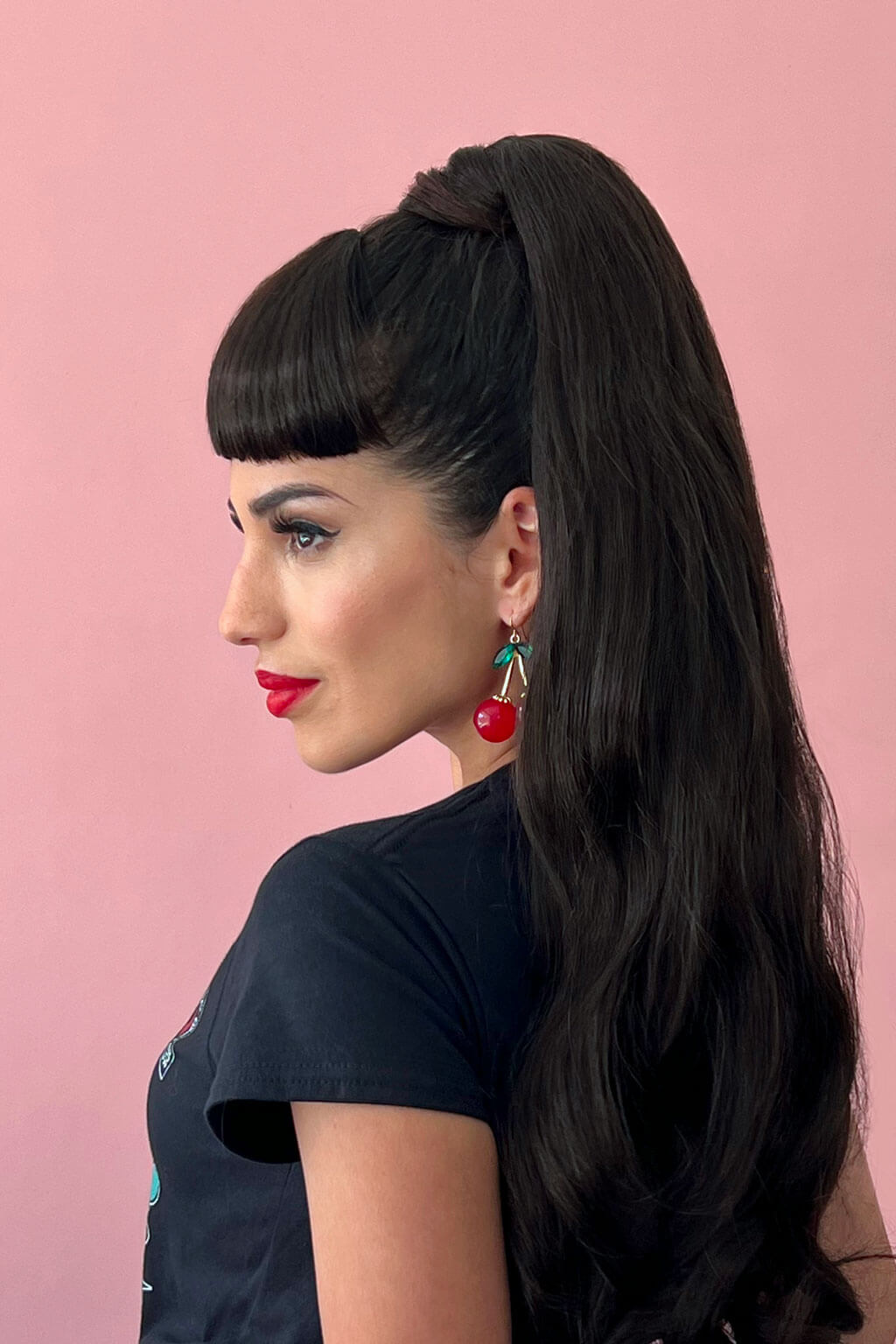 Woman with long dark hair styled in a high ponytail against a pink background. Clip-in fringe hairpiece/clip-in bangs, thick, straight retro rockabilly style: Ida dark brown 4