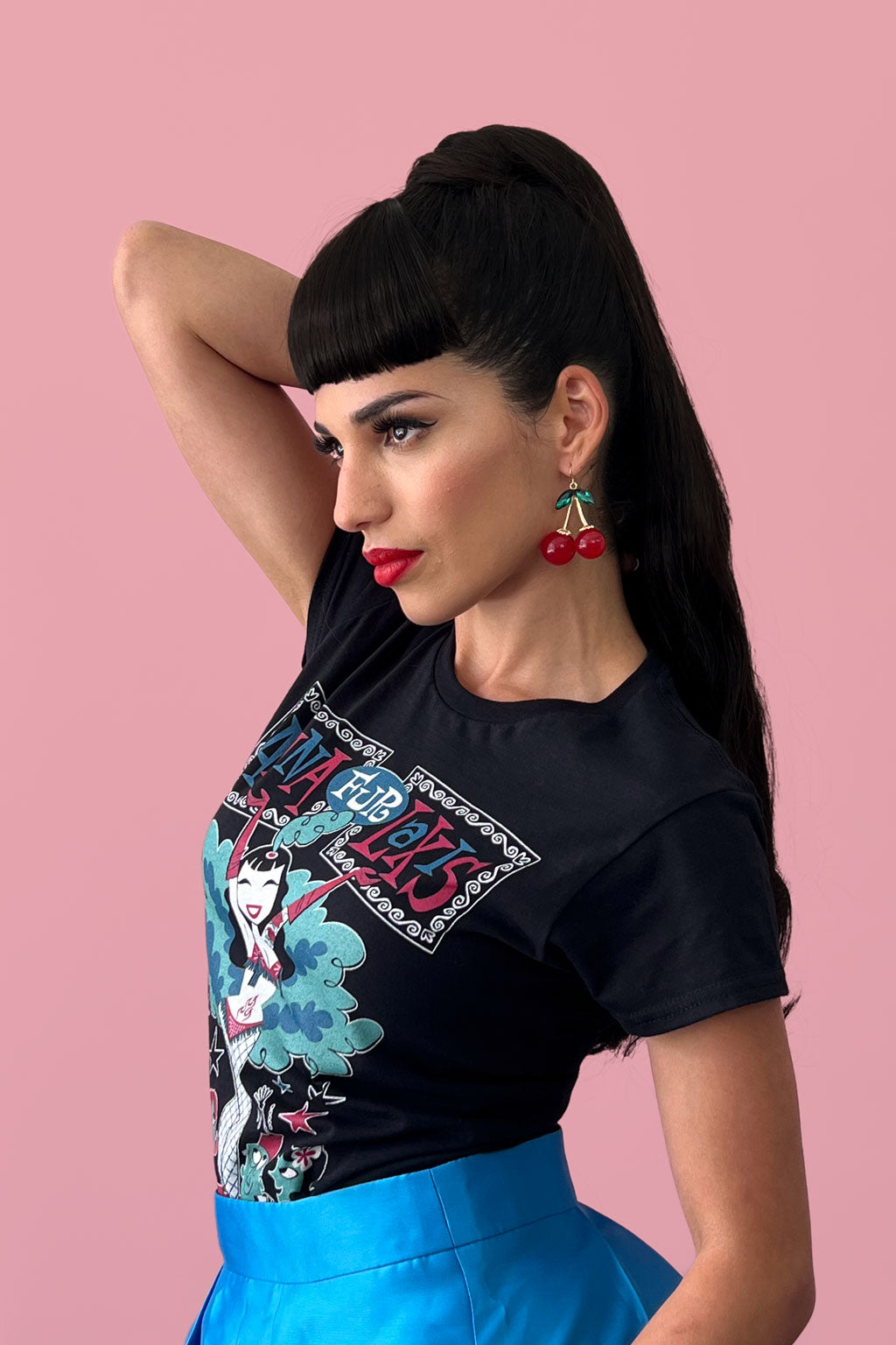 Woman wearing a black t-shirt with colorful graphics and a blue skirt against a pink background. Clip-in fringe hairpiece/clip-in bangs, thick, straight retro rockabilly style: Ida chestnut black 1B