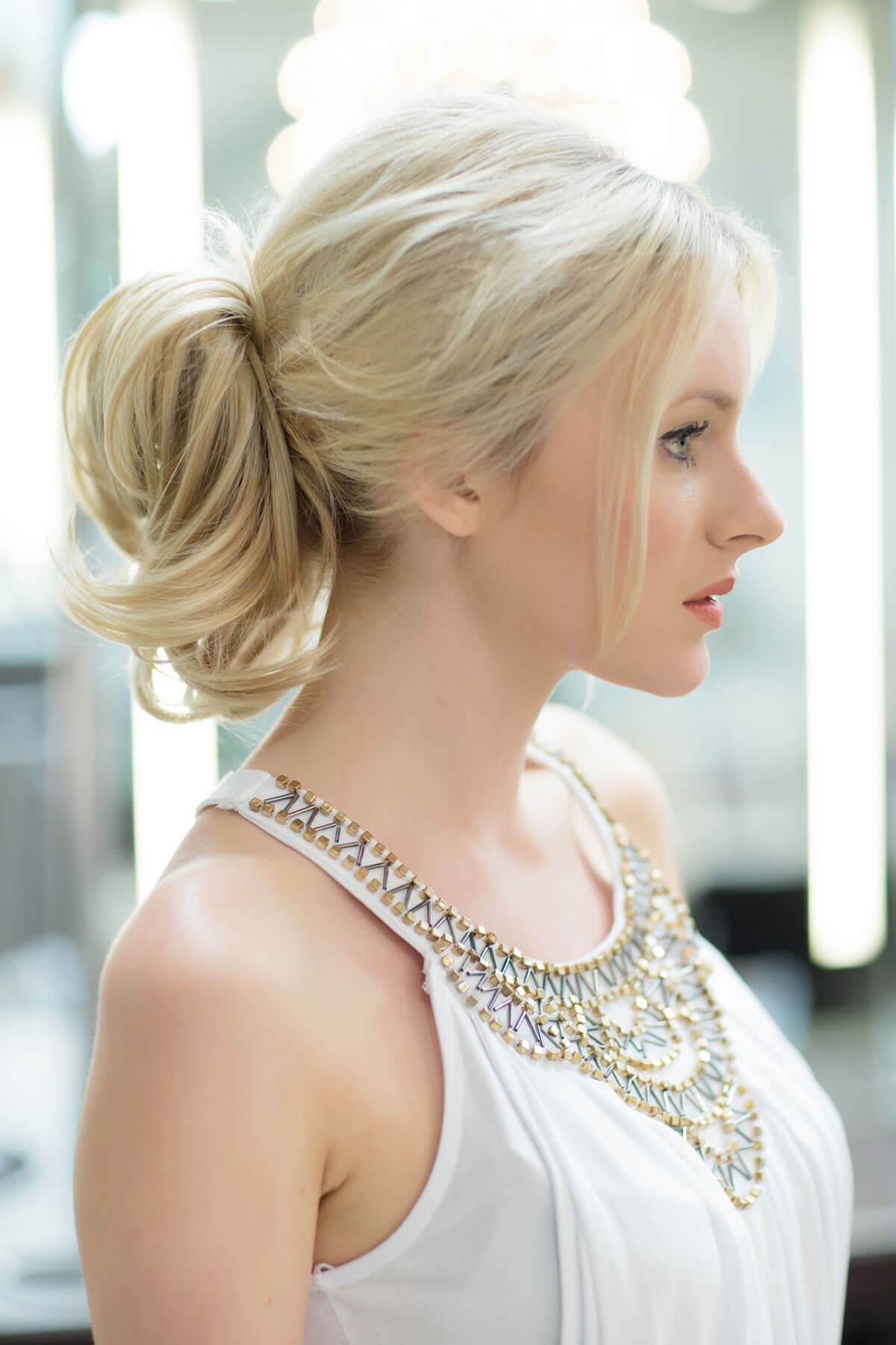 Woman with styled blonde hair wearing a white top with a decorative neckline. Clip in hairpiece, ponytail, short, flexihair: Leela champagne blonde 613
