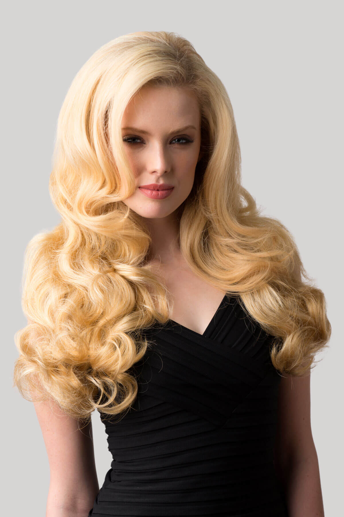 Woman with long blonde hair styled in curls against a gray background. Curly blonde half wig hairpiece extension (3/4 wig), long: Meana honey blonde 24BH613