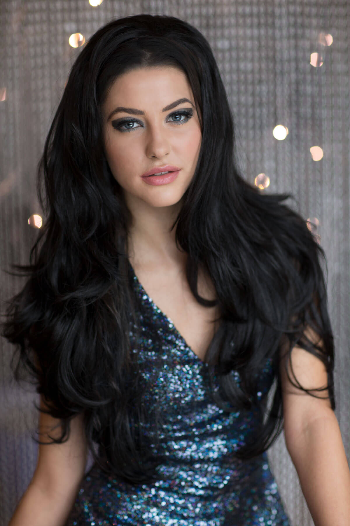 Woman with long black hair wearing a blue sequin dress against a sparkly background. Black half wig hairpiece (3/4 wig), long layers: Aislynn black #1b