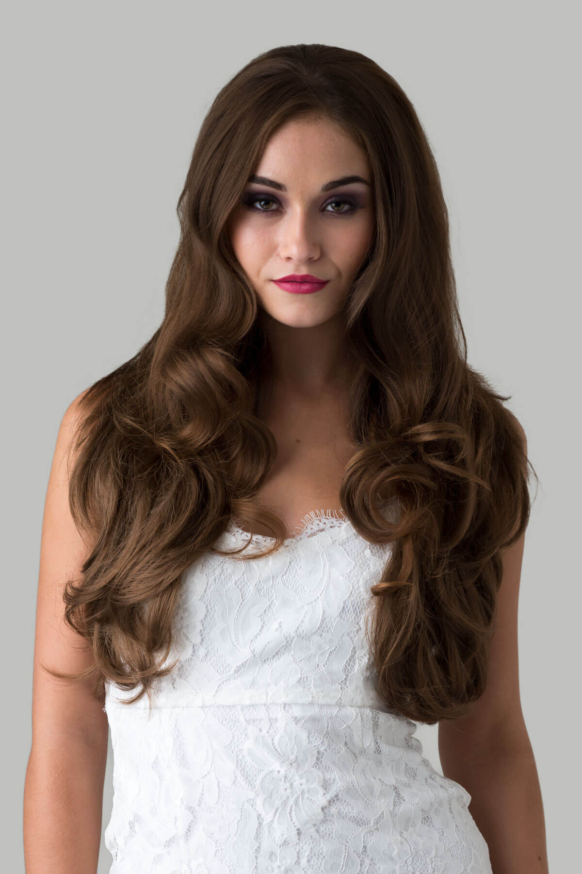 Woman with long, wavy brown hair wearing a white dress against a gray background. Brown half wig hairpiece (3/4 wig), layered, wavy: Simone light brown 12