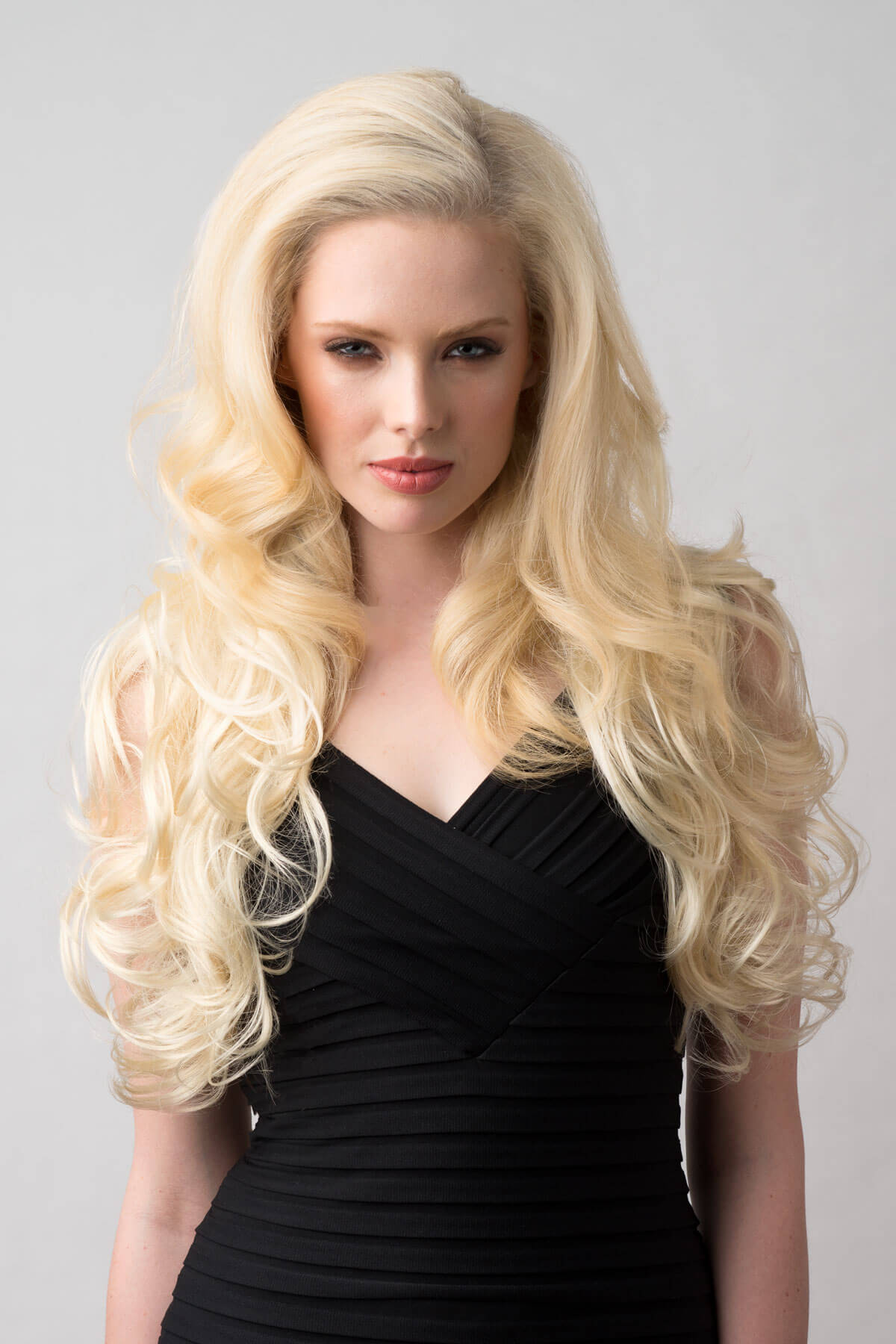 Woman with long blonde wig wearing a black dress on a gray background. Long blonde 3/4 hairpiece (half wig), big loose curls: Zara creamy blonde 613