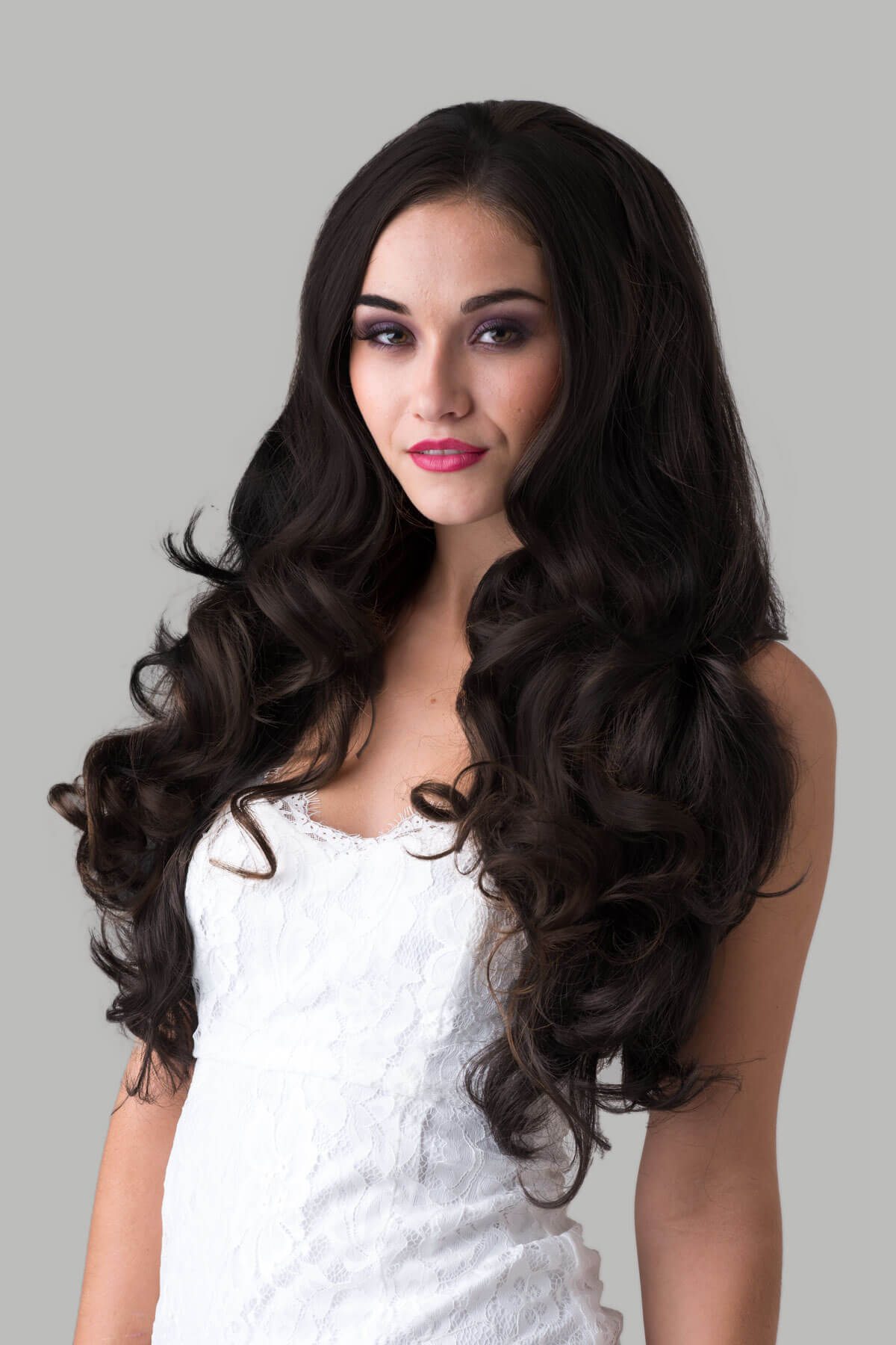 Woman with long, wavy hair wearing a white top against a gray background. Brown half wig hairpiece with long loose curls: Chelsea dark brown 4