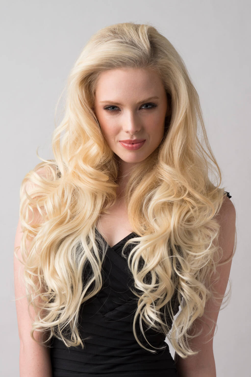 Woman with long, wavy blonde hair wearing a black top against a grey background. Blonde half wig hairpiece (3/4 wig), loose curls: Suki creamy blonde 613