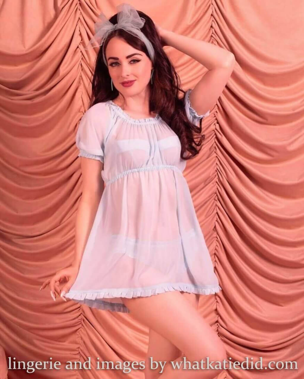 A woman in a short, sheer babydoll nightgown in pale pink and white with light blue trim, paired with a matching sheer headband, posed against a draped peach-pink fabric backdrop. High crown long, wavy half wig and volume ring: Bardot chestnut brown 8