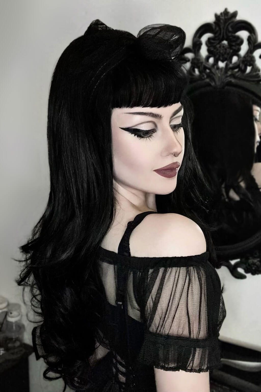 Woman with long black hair and dark makeup wearing a black outfit. Woman wearing a pink top with black patterns and a black skirt, standing indoors. High crown long, wavy half wig and volume ring: Bardot jet black 1