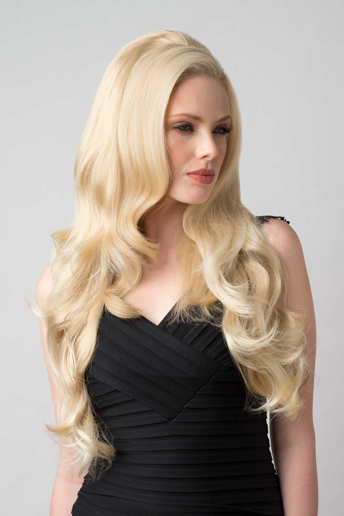Woman with long blonde wig wearing a black dress on a gray background. Half wig with volume booster/height booster: Corrine blonde 613T24