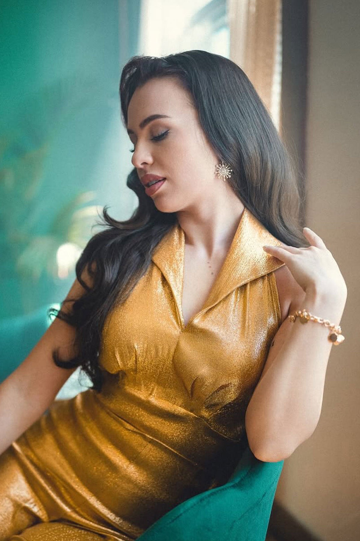 Woman in a gold dress sitting on a green chair with a blurred background. Brown half wig hairpiece, full, long with gentle waves: Taya chestnut brown 8