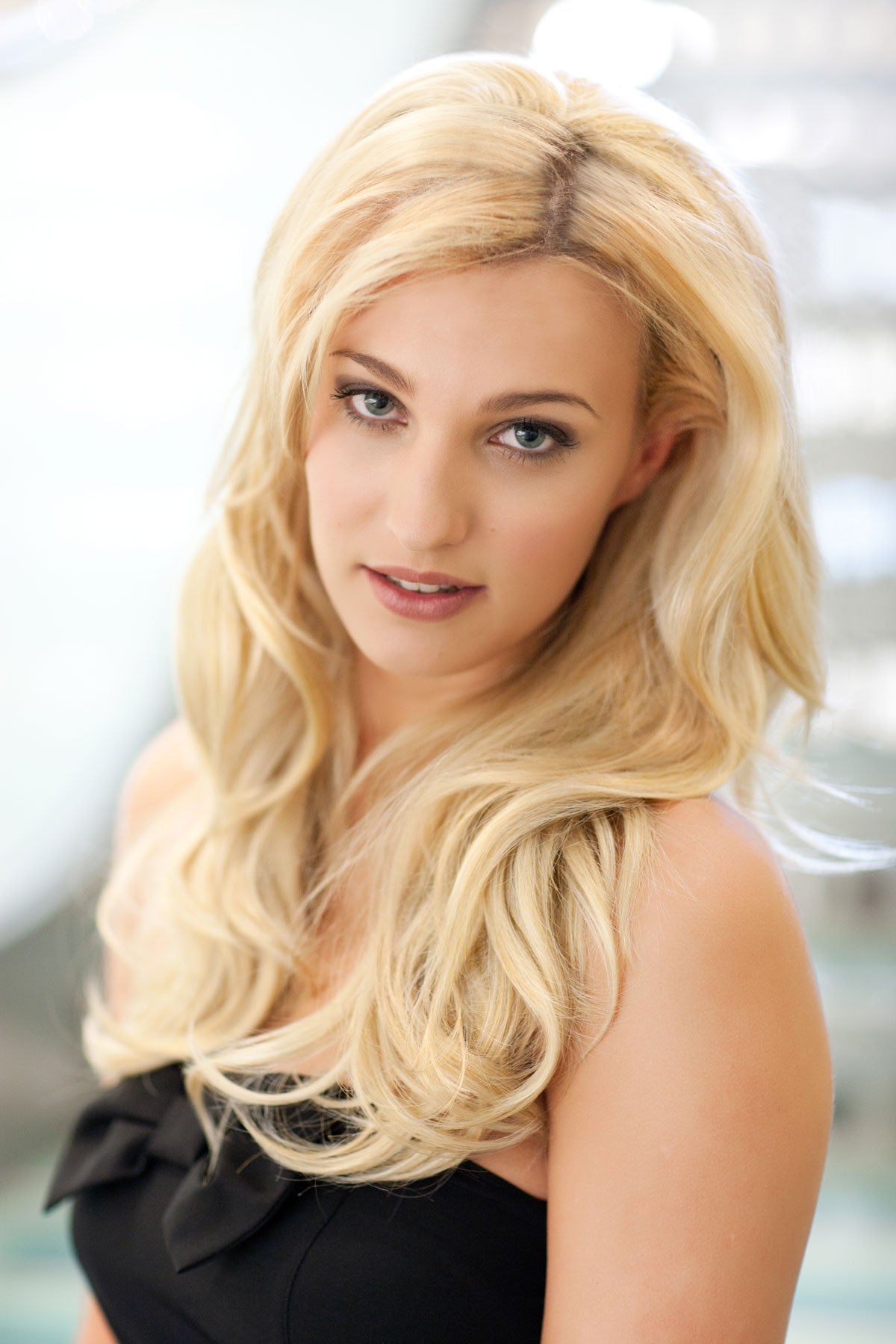 Woman with long blonde hair wearing a black top against a blurred background. Heat styleable half wig hairpiece (3/4 wig), long, wavy, Flexihair: Michela blonde 24BH613