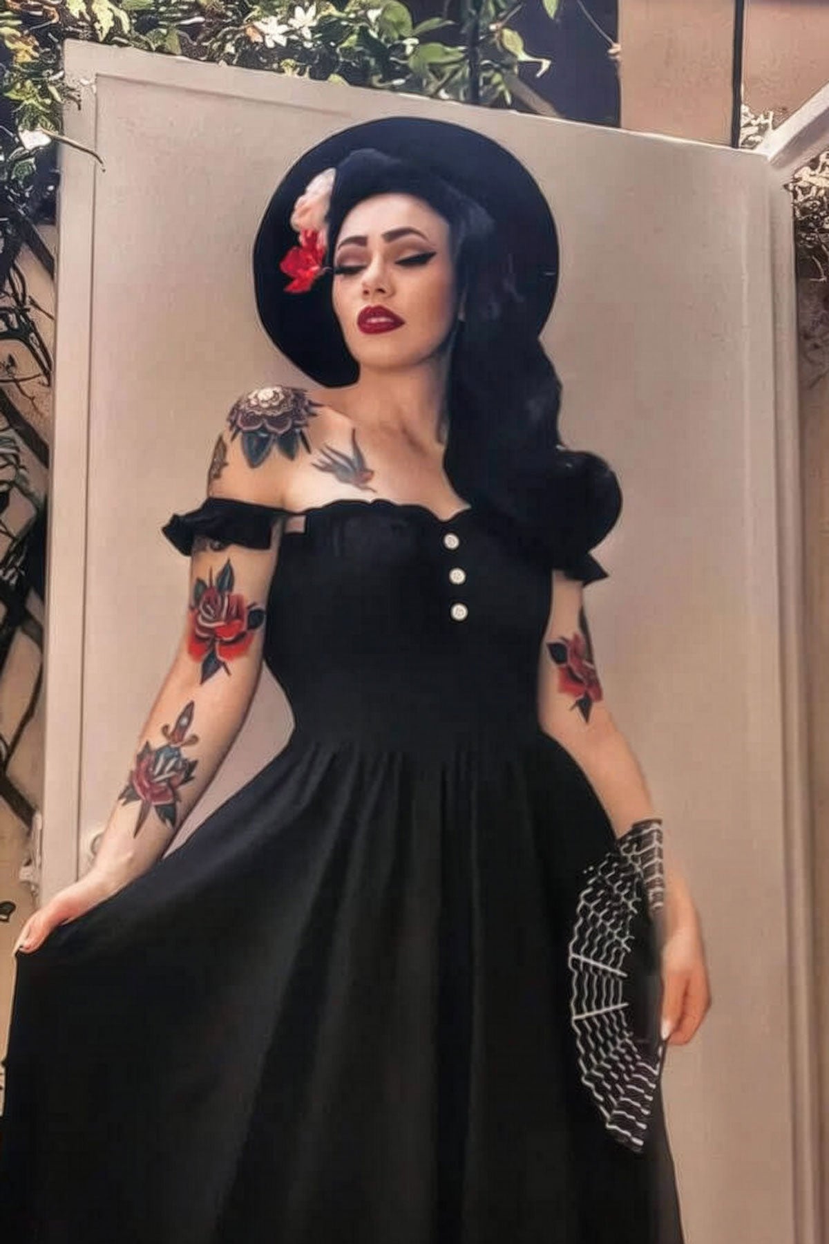 Woman in a black dress and hat with tattoos standing against a neutral background. 1960s Retro high crown, half wig hairpiece, long with hair flip: Priscilla black 1B