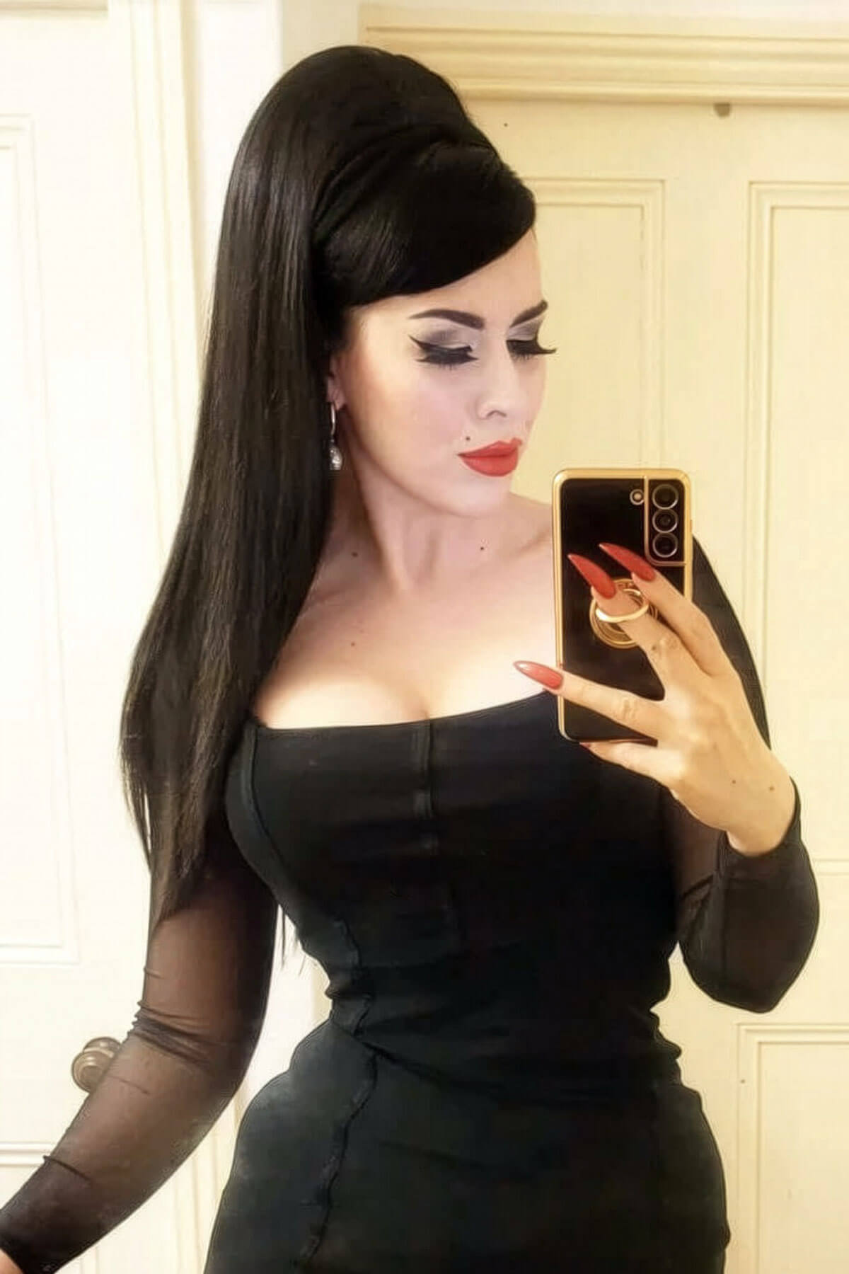 Woman in a black dress taking a mirror selfie with a phone. 1960s style hair, high crown long straight half wig: Andrea black 1B