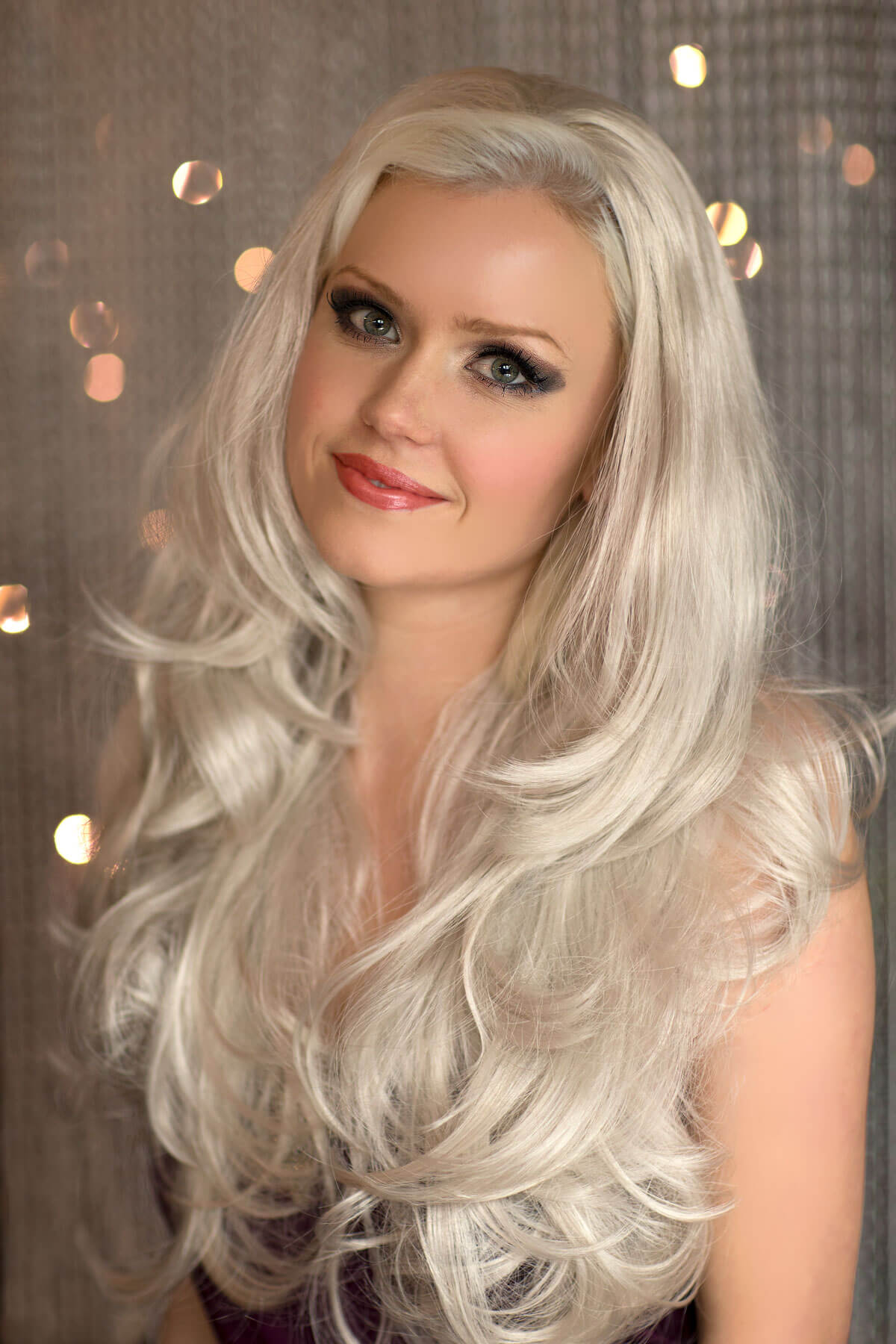 Woman with long, wavy silver hair against a blurred background with lights. Silver half wig hairpiece (3/4 wig), loose curls: Tahlia