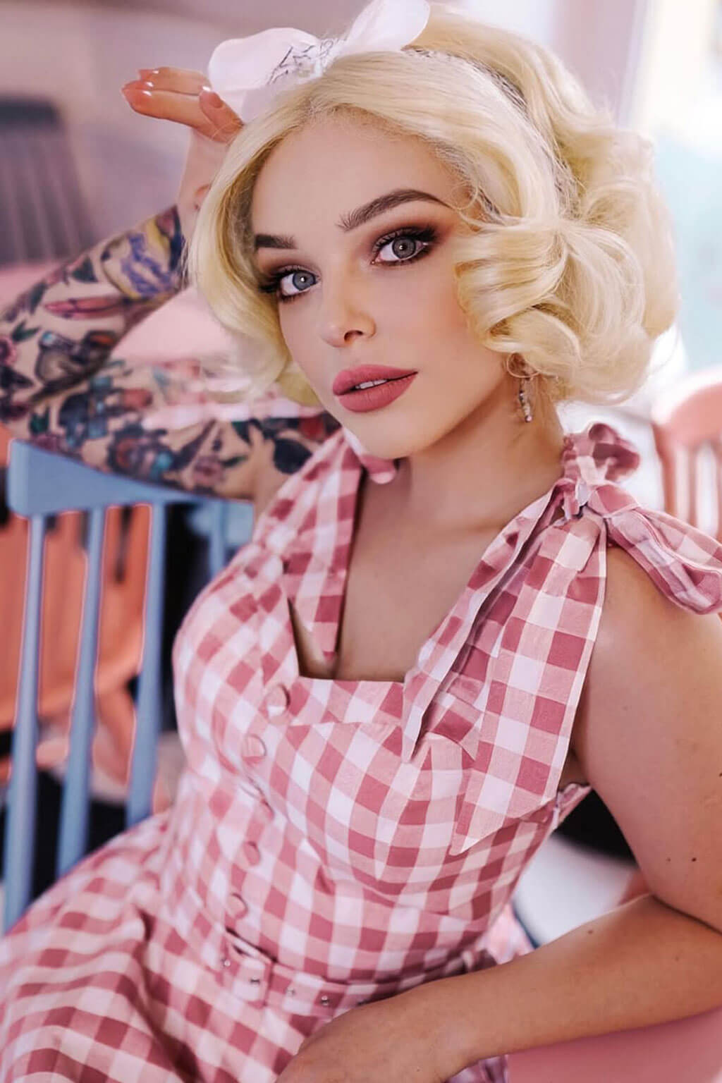 Woman in a pink checkered dress with styled hair, sitting on a chair. Blonde pinup wig, lace front, vintage style: Marilyn creamy blonde 613