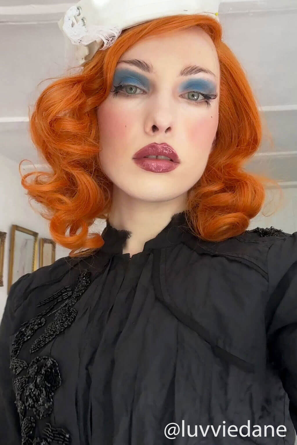 Person with vibrant orange hair and dramatic makeup wearing a black outfit. Ginger lacefront wig, pinup/vintage style, mid length with finger waves: Flora