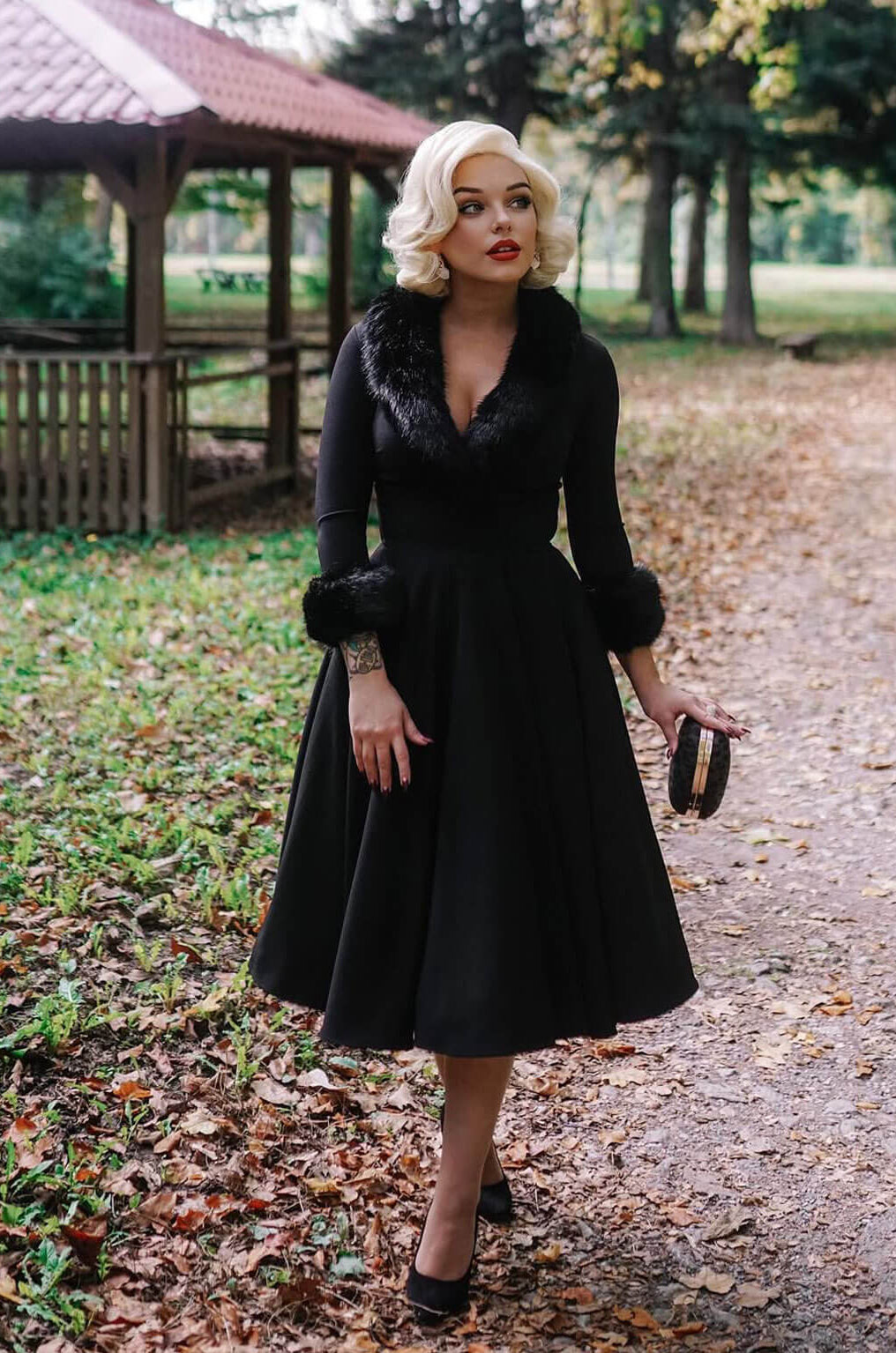 Woman in a black dress with fur trim standing on a path in a park. Blonde pinup wig, lace front, vintage style: Marilyn white blonde 1001