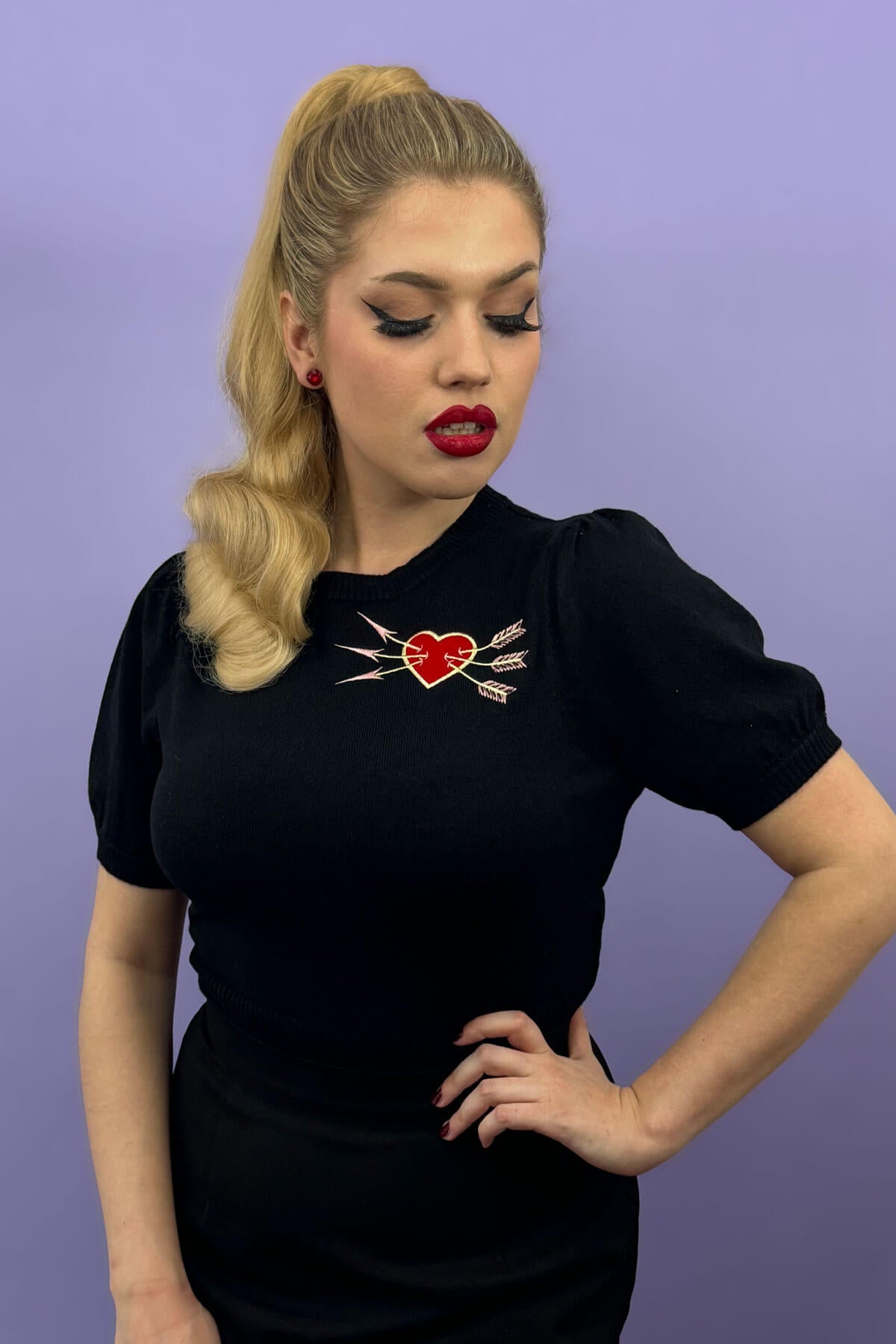Woman wearing a black t-shirt with a red heart and arrow design against a purple background. Retro style ponytail hairpiece, long with vintage waves: Arabella blonde 24H613