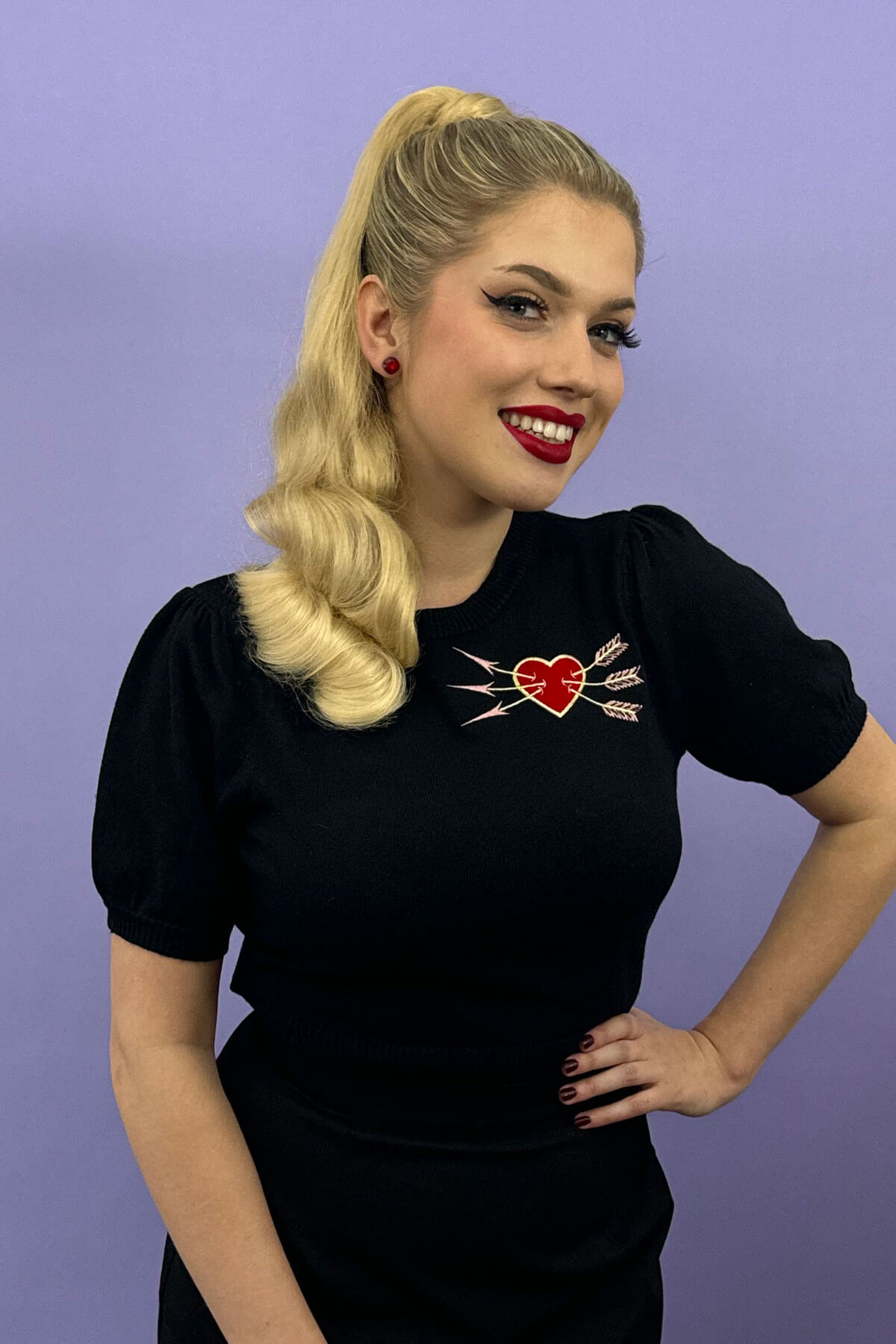 Woman wearing a black dress with a heart design on a purple background. Retro style ponytail hairpiece, long with vintage waves: Arabella champagne blonde 613