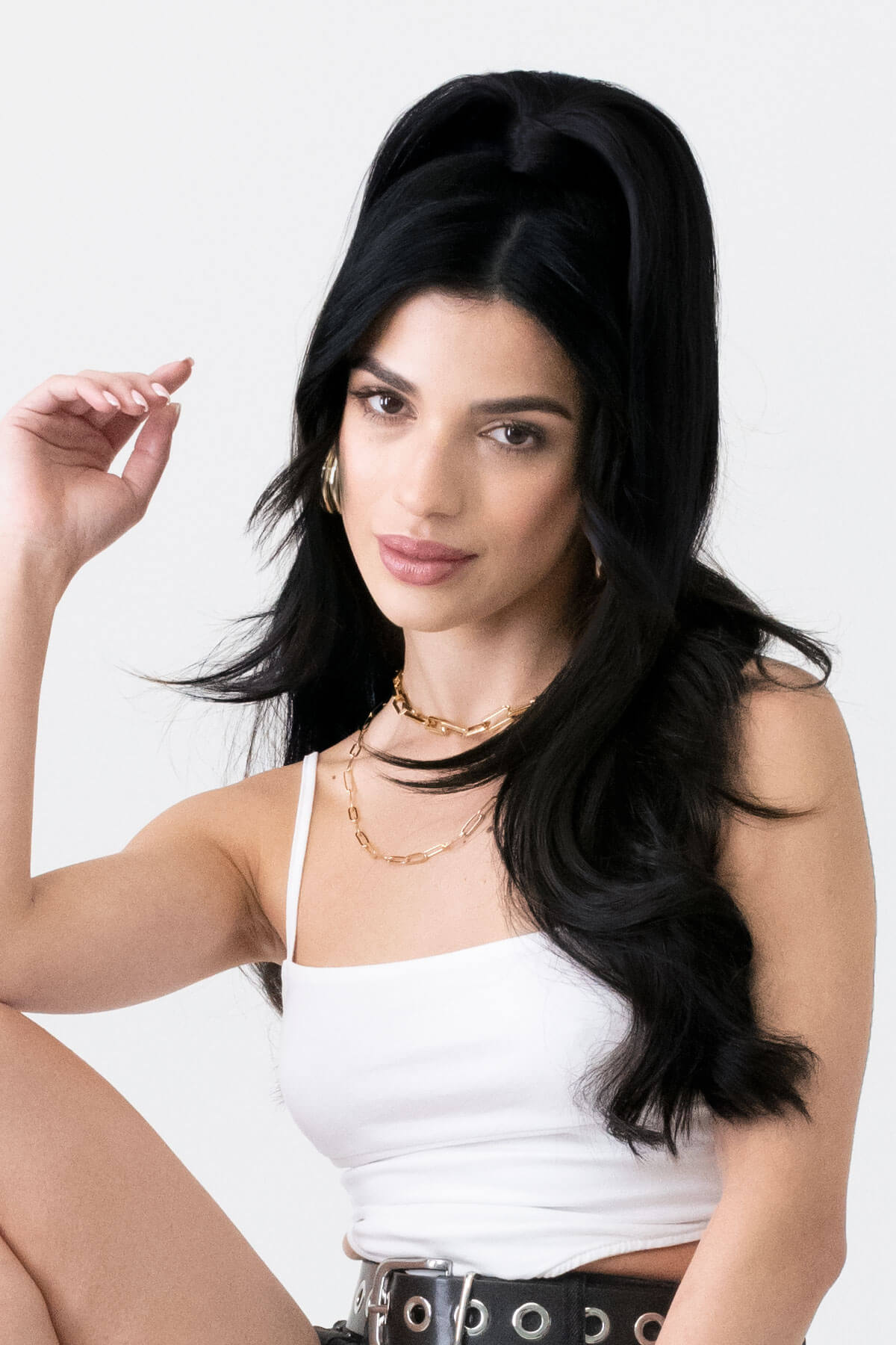Woman with long black hair wearing a white top and gold necklaces on a white background. Long sleek ponytail extension with gentle, layered waves: Mali black 1b
