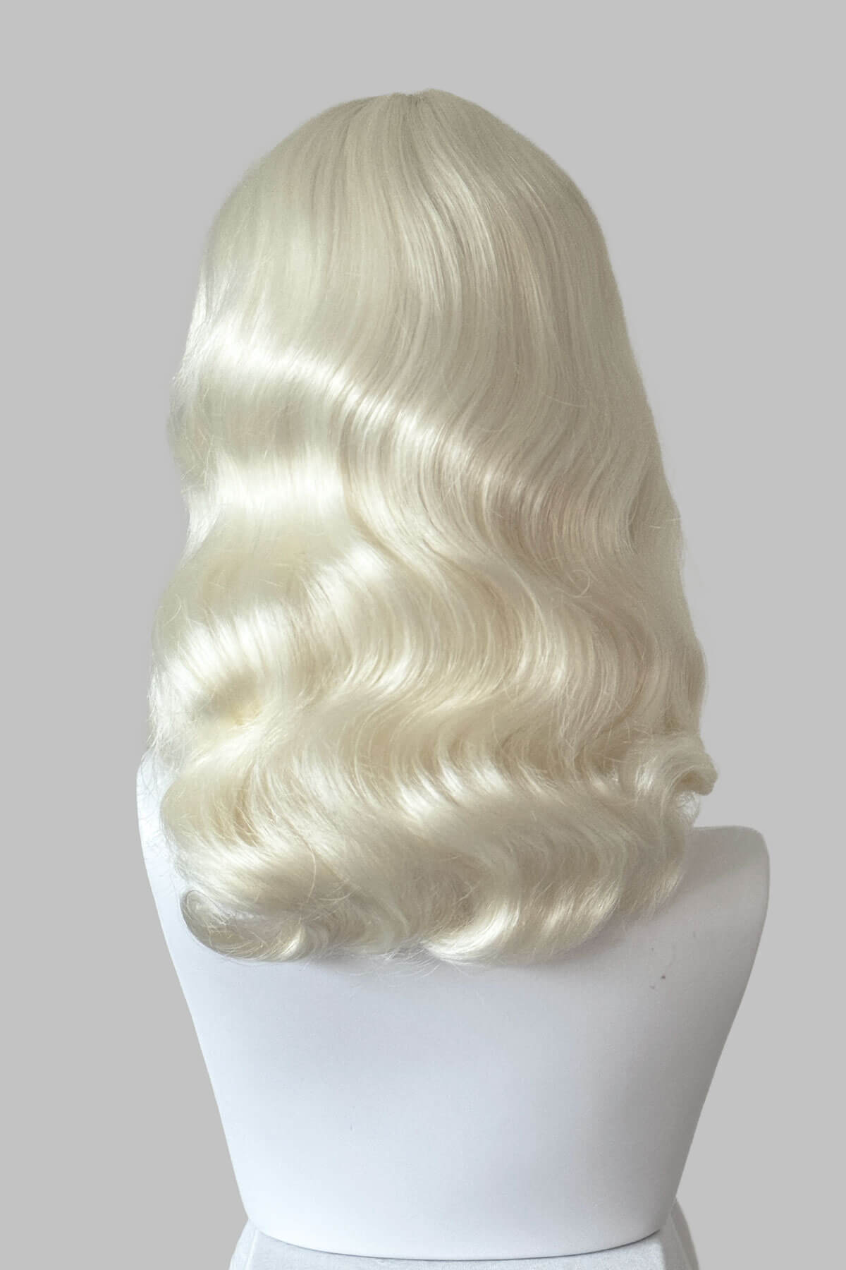 Blonde wig on a mannequin head against a plain background. Platinum blonde pin-up style wig with short fringe and marcel waves: Eira