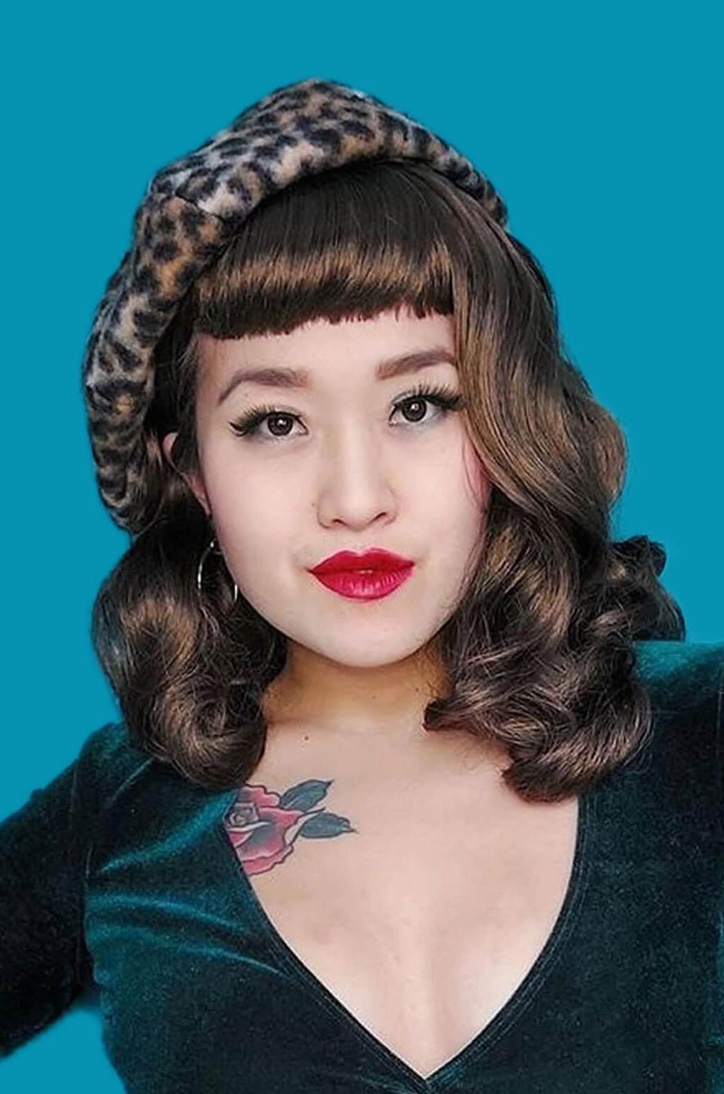 Woman wearing a leopard print hat and green velvet top against a blue background. Brown pinup style wig, curled with short fringe: Carolina mocha brown 10