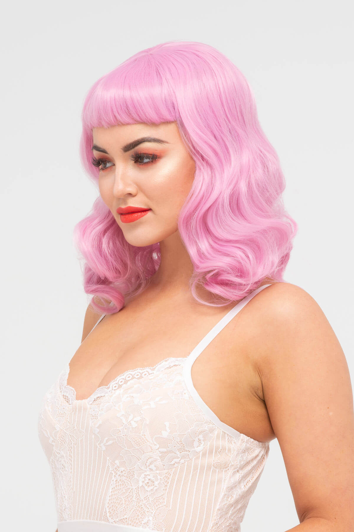 Woman with pink wig and red lipstick wearing a white lace top on a light gray background. Pink pinup style wig, with finger waves and a short fringe, 1950s style: Stevie light pink 2614