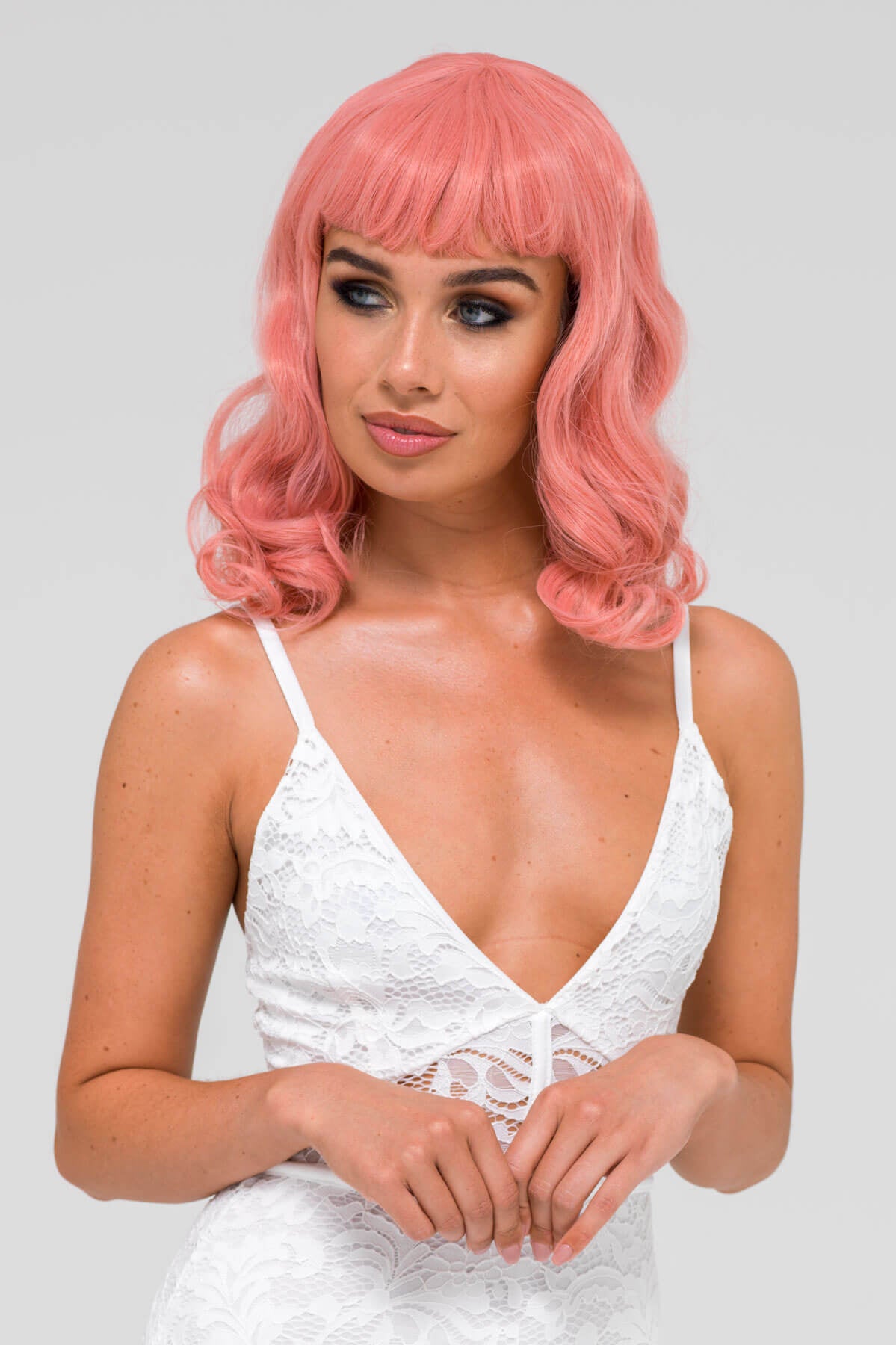 Woman with pink wig wearing a white lace dress on a light gray background. Pink pinup style wig, with finger waves and a short fringe, 1950s style: Stevie