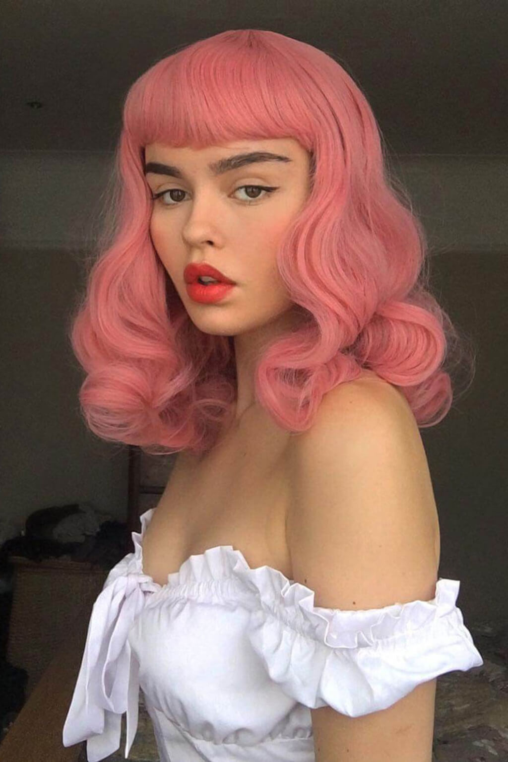 Person with pink hair and red lipstick wearing a white off-shoulder top. Pink pinup style wig, with finger waves and a short fringe, 1950s style: Stevie