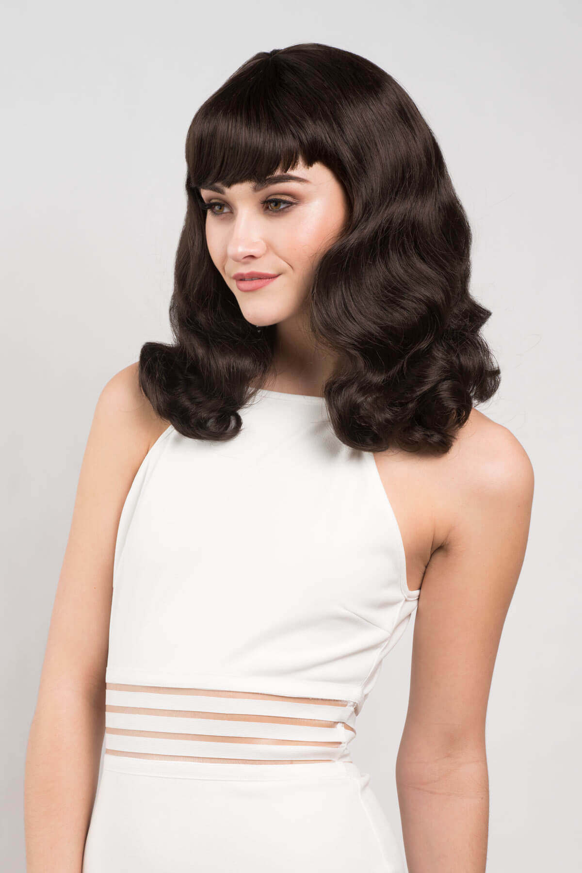 Woman with vintage-style hair wearing a white dress against a light gray background. Dark brown pinup style wig, finger waved with short fringe, 1950s style: Becca