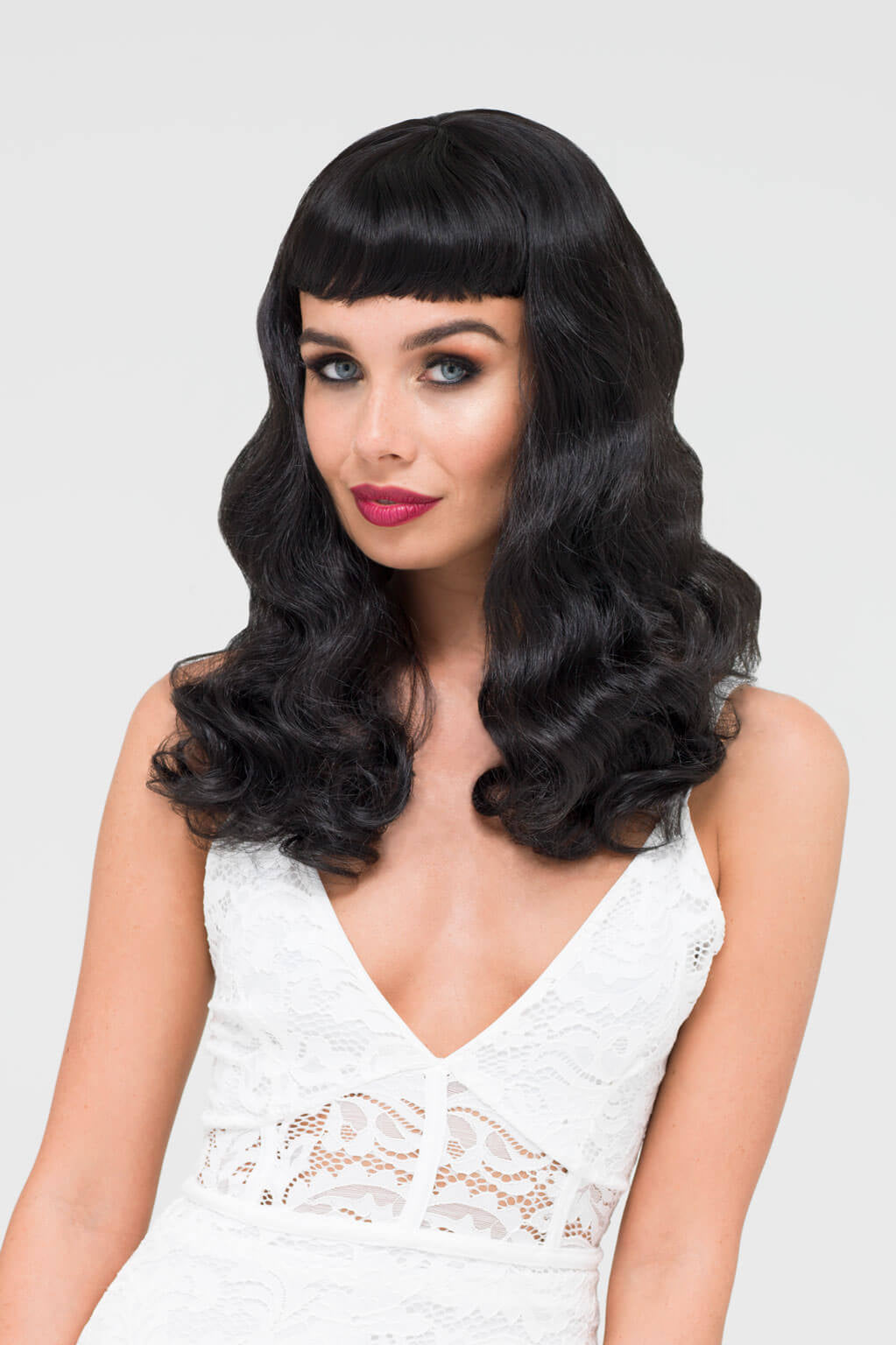 Woman with long black wavy hair and bangs wearing a white lace top on a light gray background - Black 1950s pinup style wig, curled with short fringe: Milena
