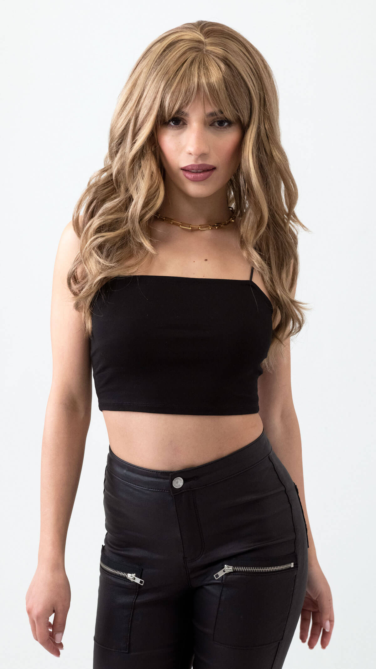 Woman wearing a black crop top and black pants with zippers on a white background. Long brown wig with blonde highlights and gentle waves: Darcy