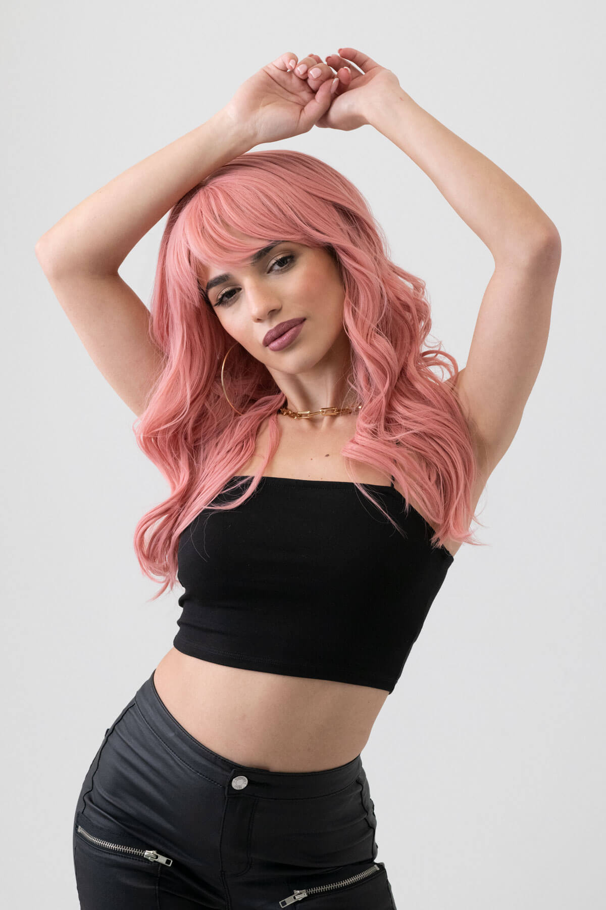 Woman with pink hair wearing a black top and black pants on a white background. Dusty pastel pink wig with long cascading waves: Rosalie