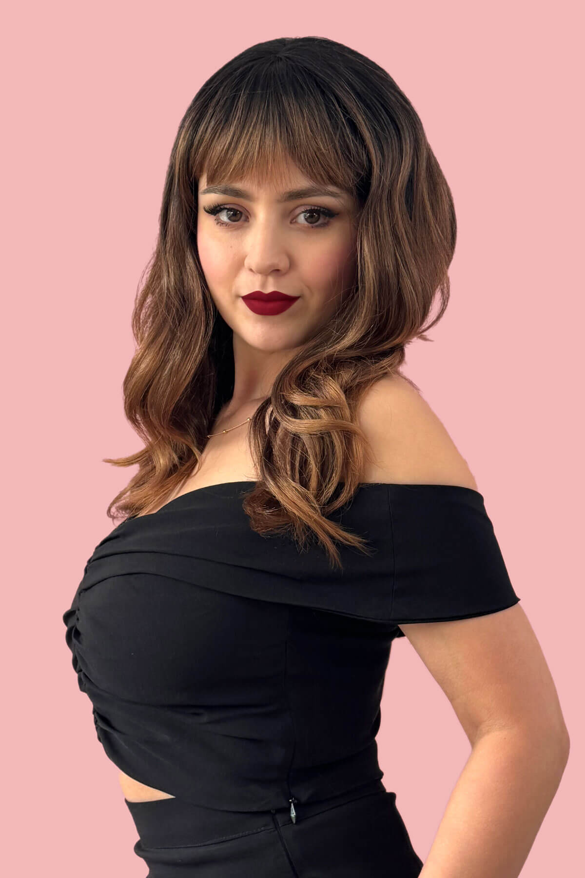 Woman wearing a black off-shoulder top against a pink background. Ombre brown wig with reddish brown highlights & waves: Serenity