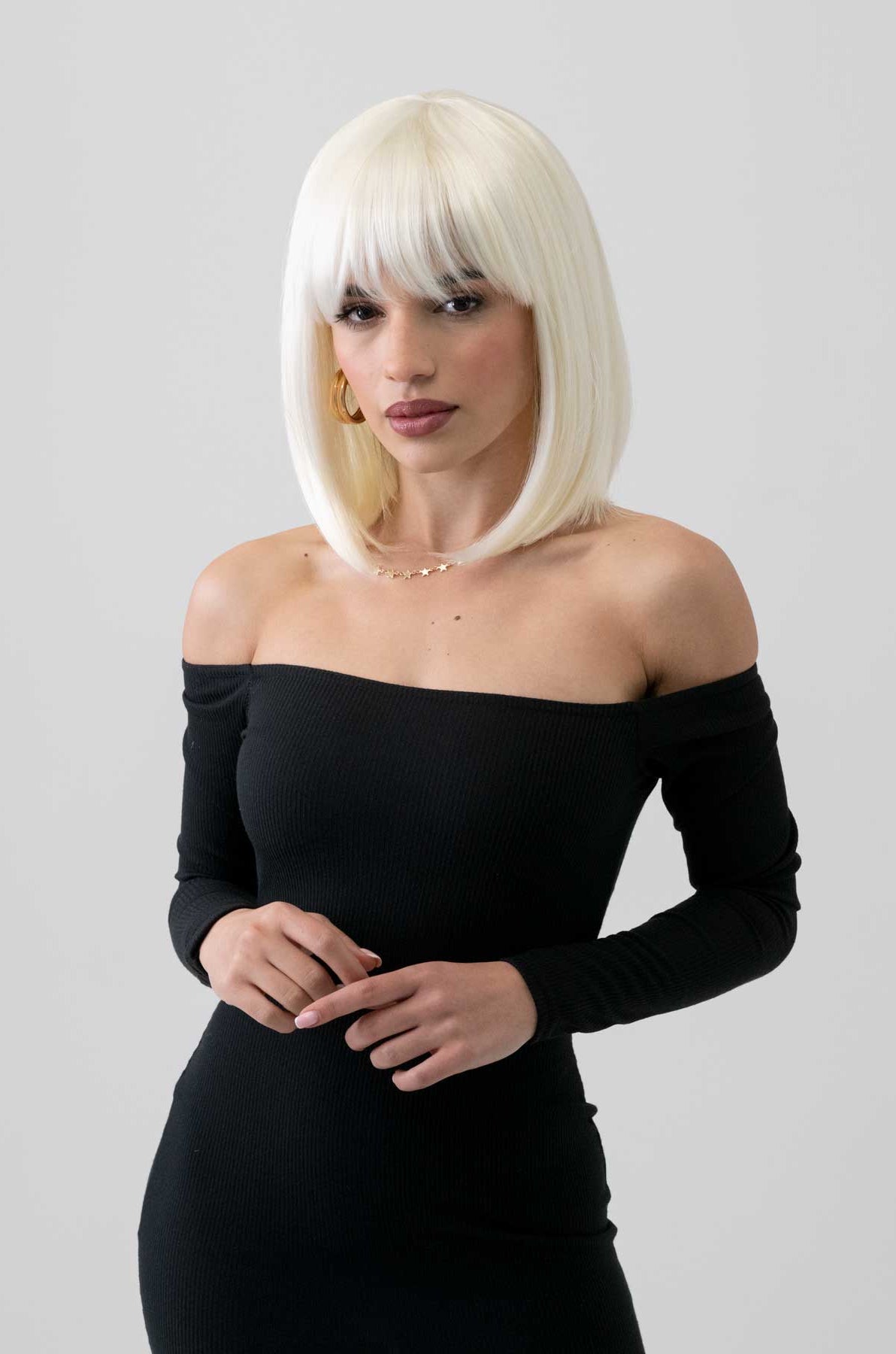 Woman with platinum blonde hair wearing a black off-shoulder dress with a plain background. Platinum blonde bob wig, shoulder length: Neve