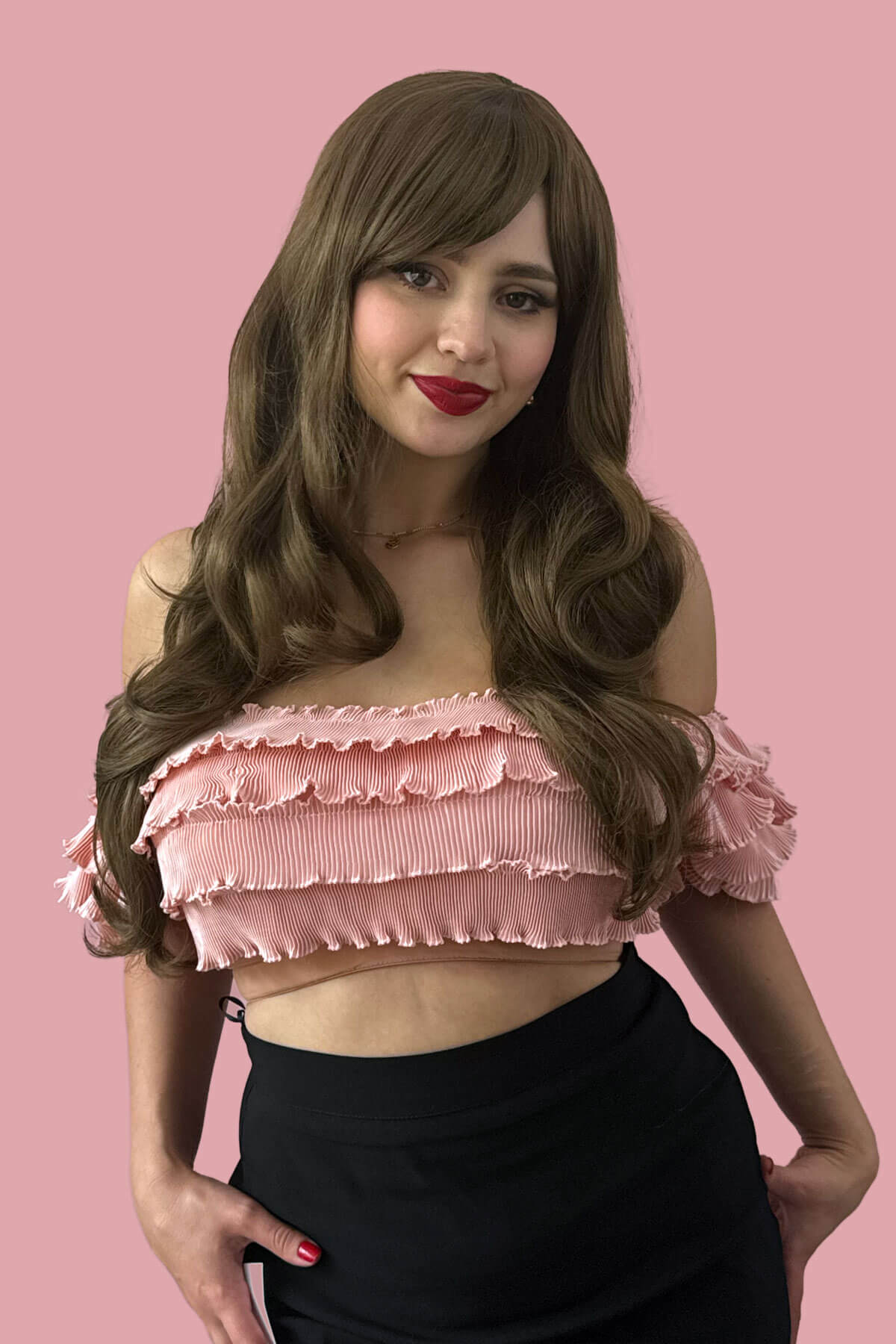 Woman wearing a pink ruffled top against a pink background. Long wavy light brown wig with vintage-style straight fringe: Maya