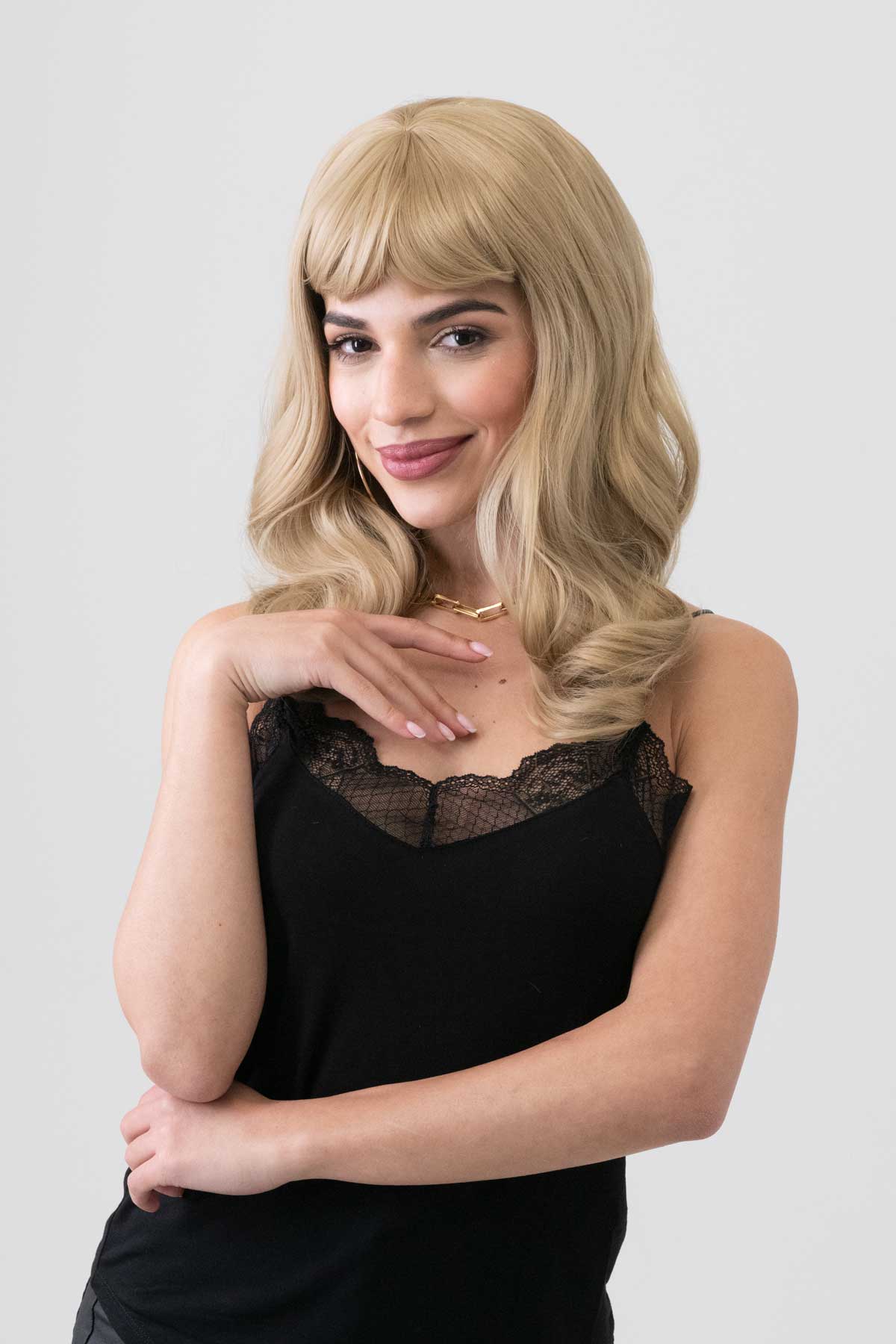 Woman with blonde wig wearing a black top against a plain background. Ash blonde 50s style wig with short fringe and gentle waves: Trinity