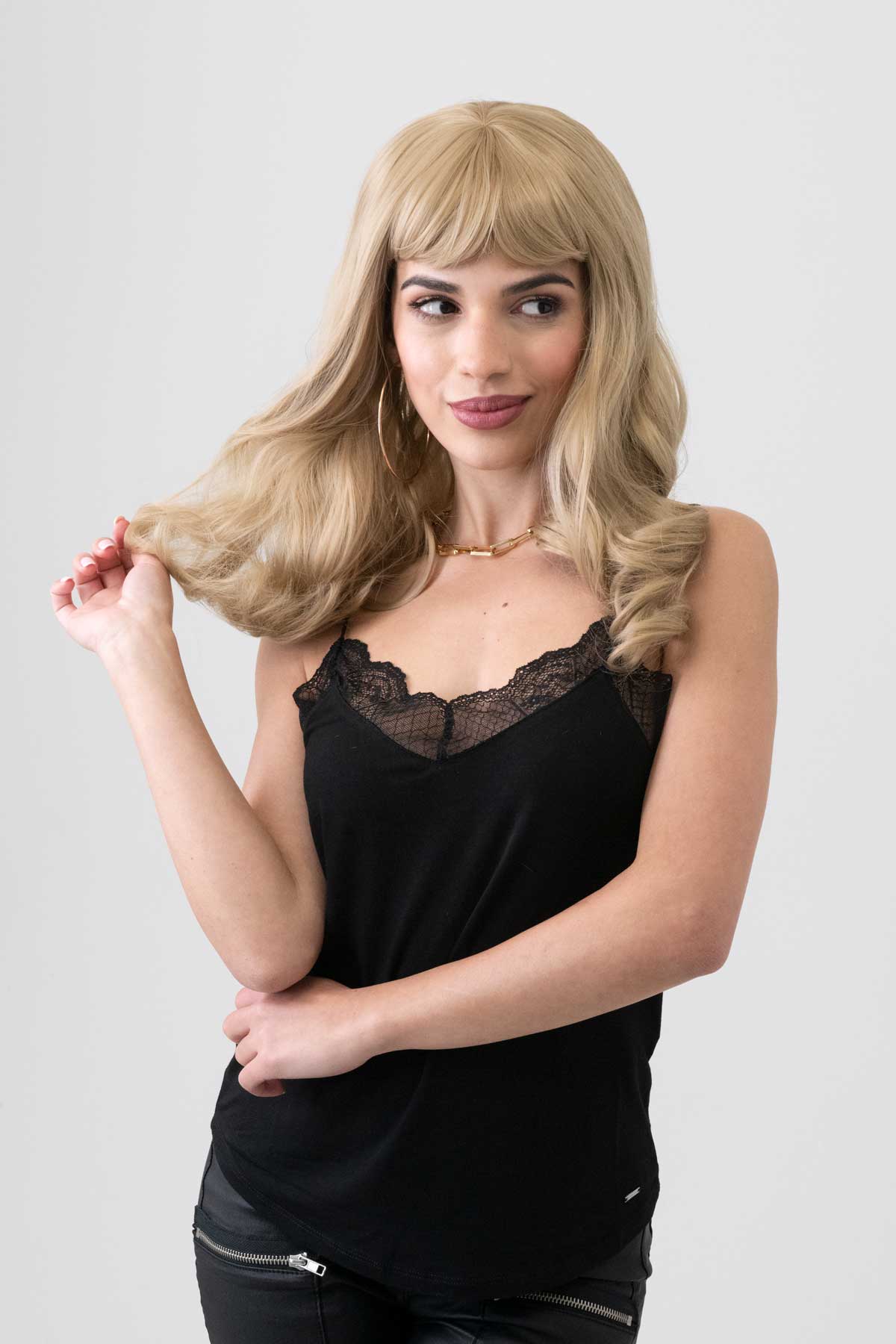 Woman wearing a black sleeveless top with lace detail on a plain background. Ash blonde 50s style wig with short fringe and gentle waves: Trinity