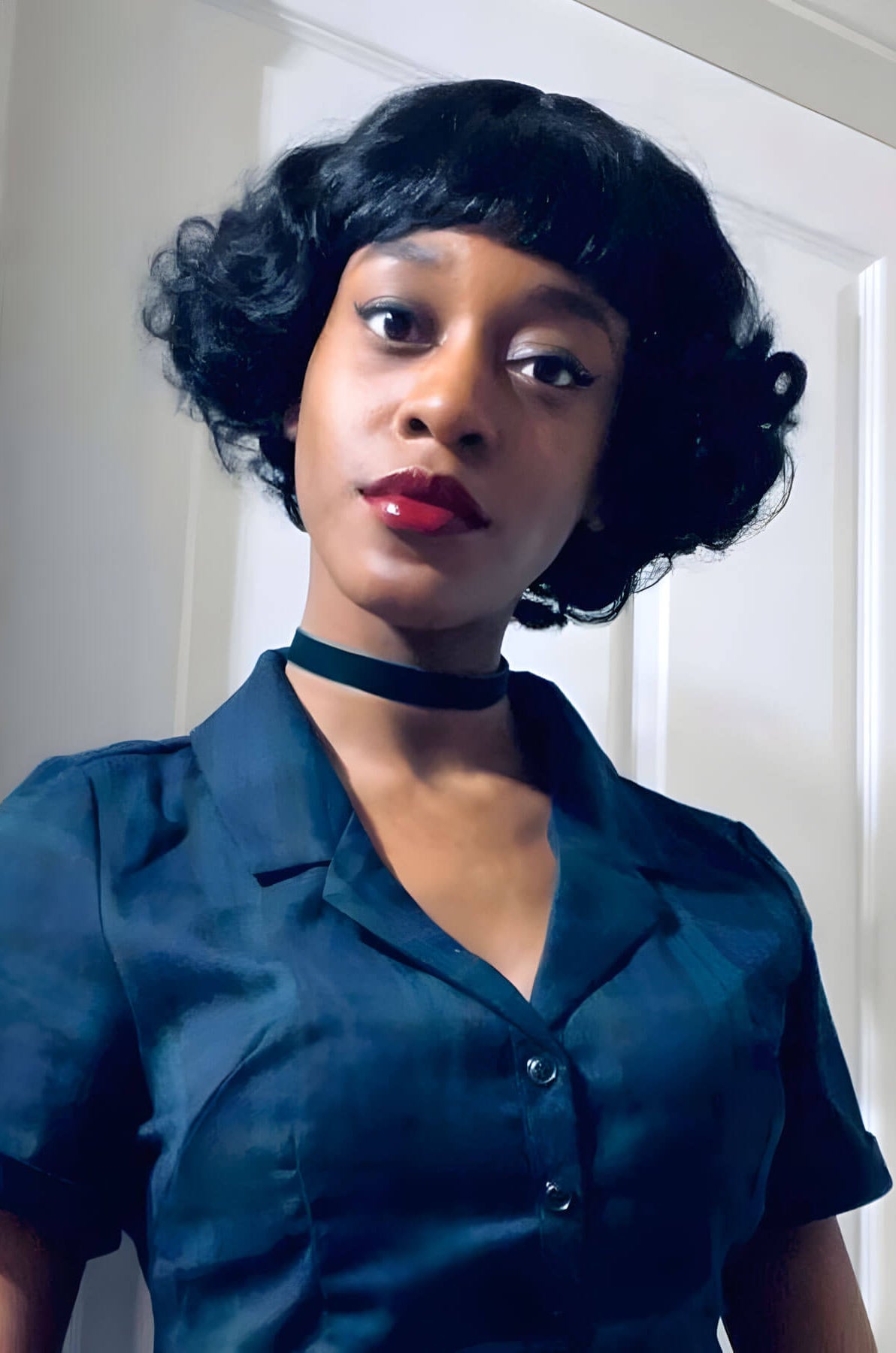 Woman wearing a teal dress with a black choker in front of a white door. A short black vintage style wig with finger waves: Marlene