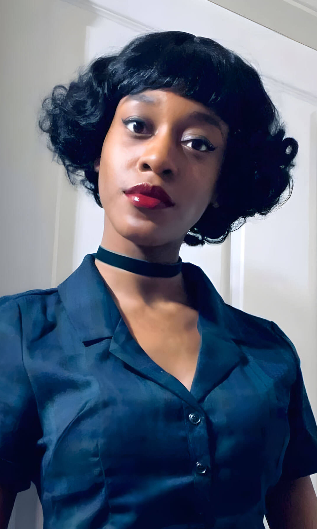 Woman wearing a teal dress with a black choker in front of a white door. A short black vintage style wig with finger waves: Marlene