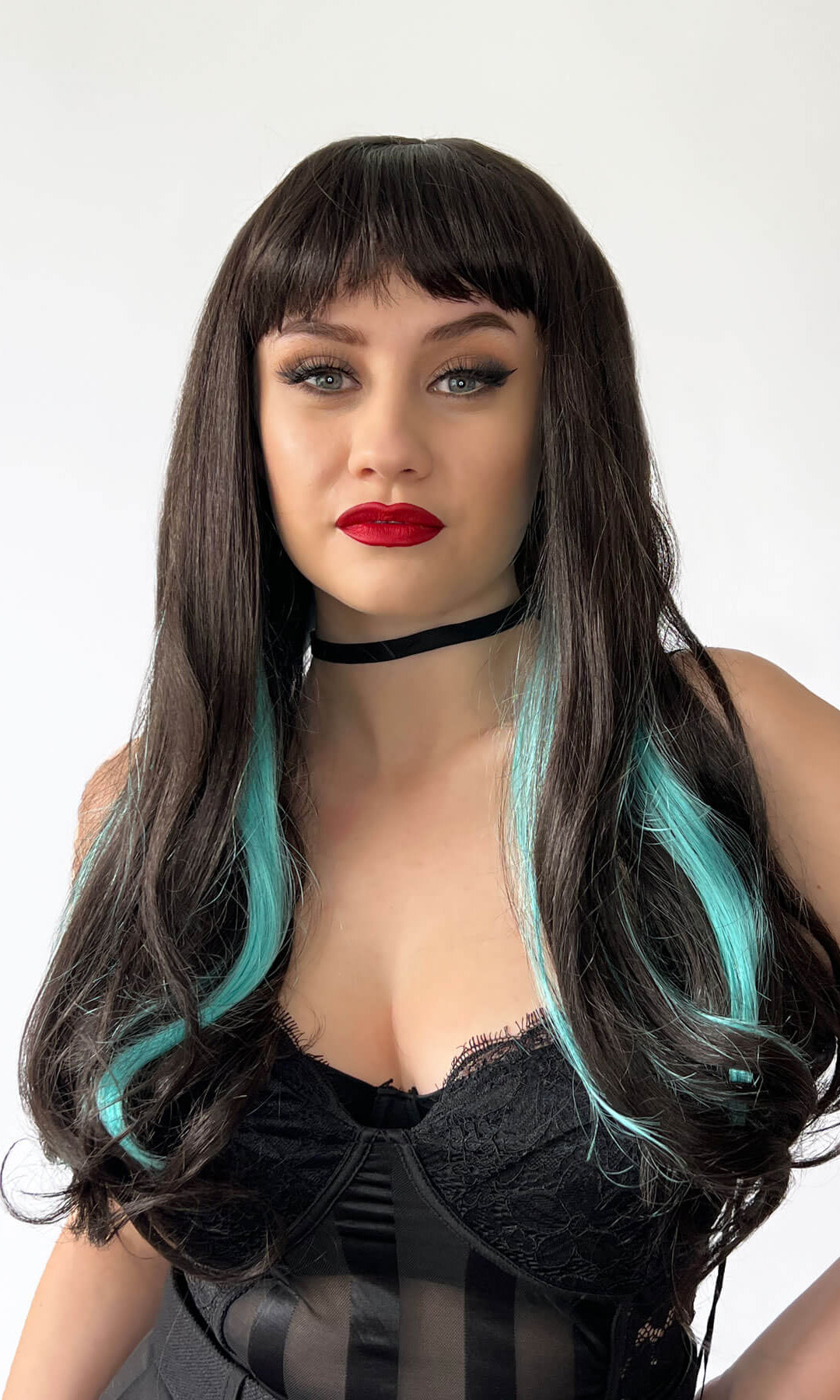 Woman with a black wig with teal highlights wearing a black outfit on a white background. Long dark brown wig with blue streaks and a straight fringe: Cyane